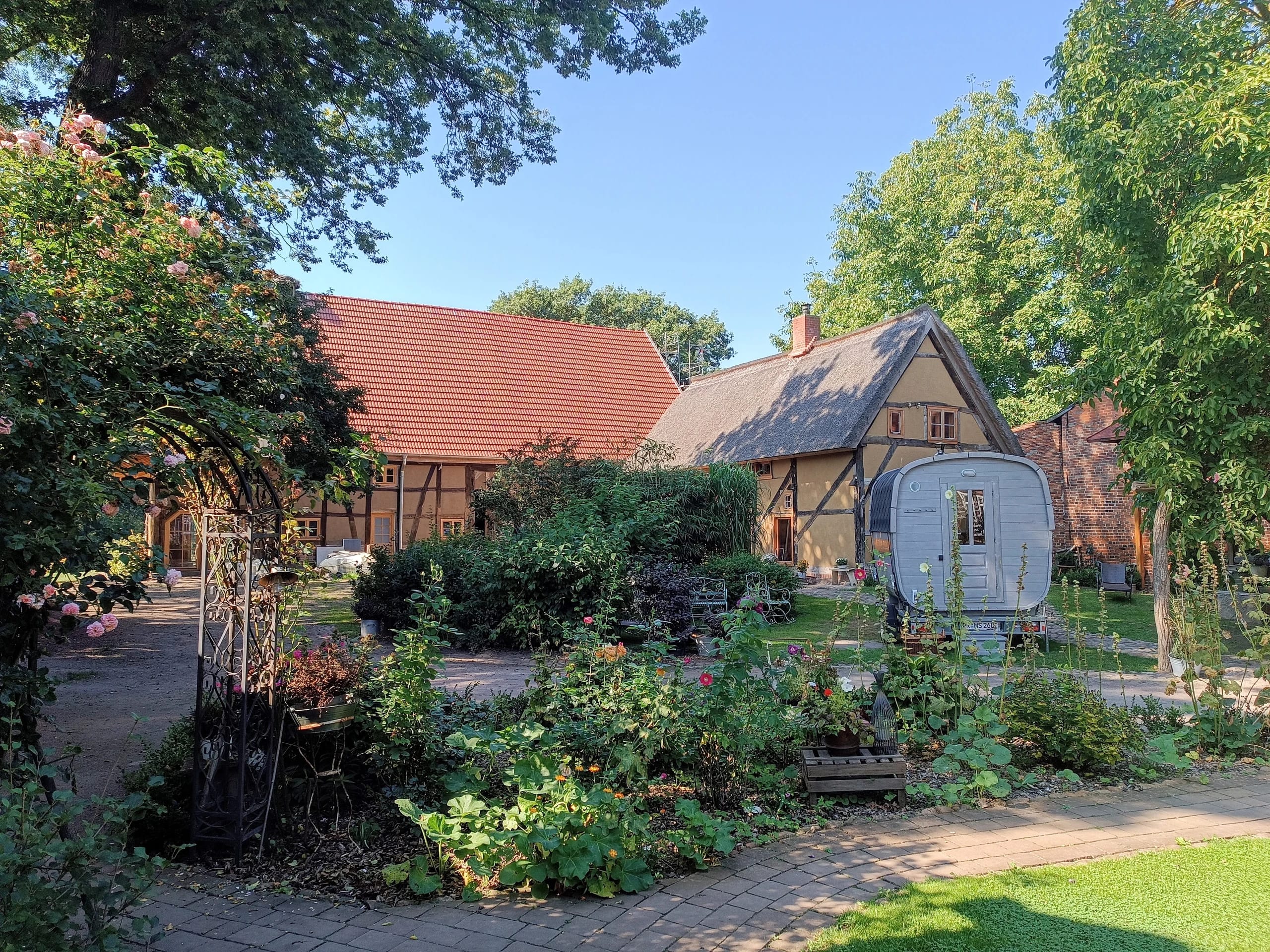 Sommerlicher Gartenhof mit Blumenbeeten, Rosenbogen und Fasssauna vor den Fachwerkgebäuden