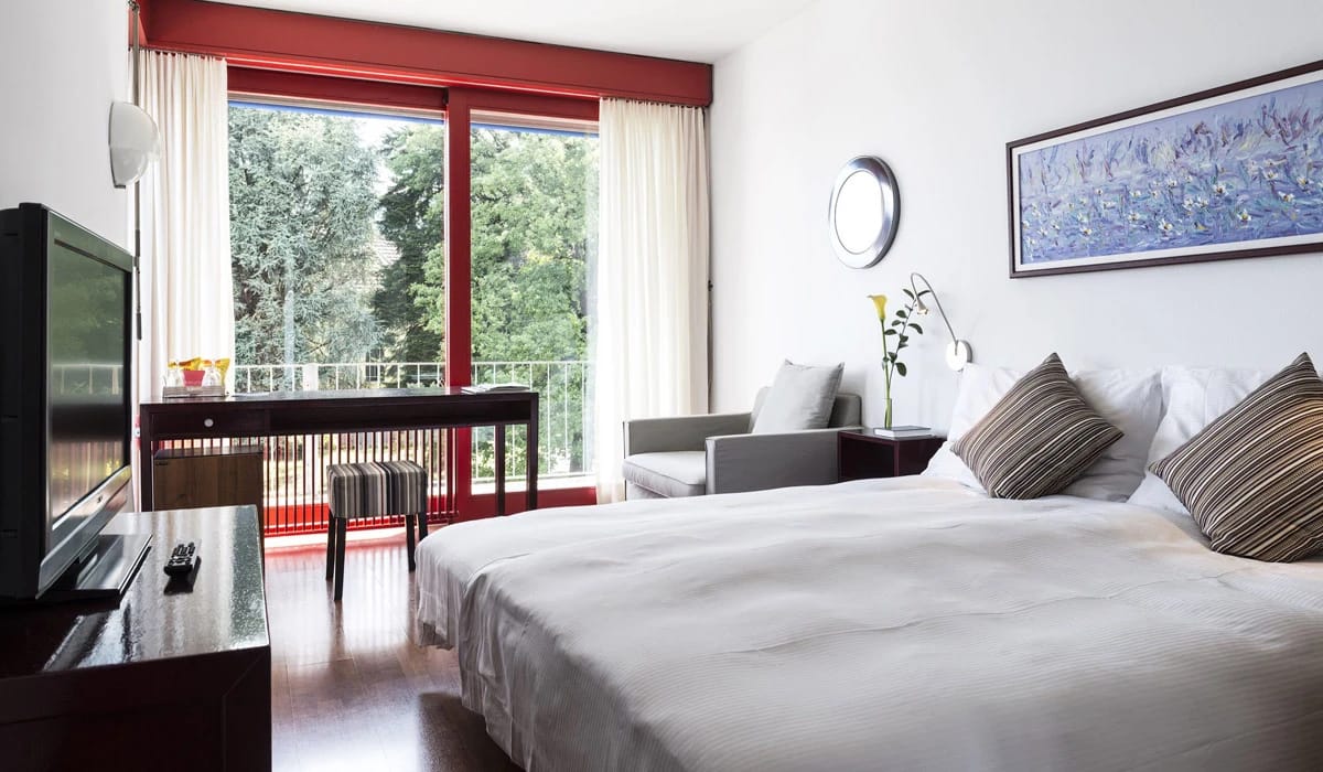 Zimmer mit Doppelbett, Sessel und rot gerahmter Balkontür mit Blick ins Grüne