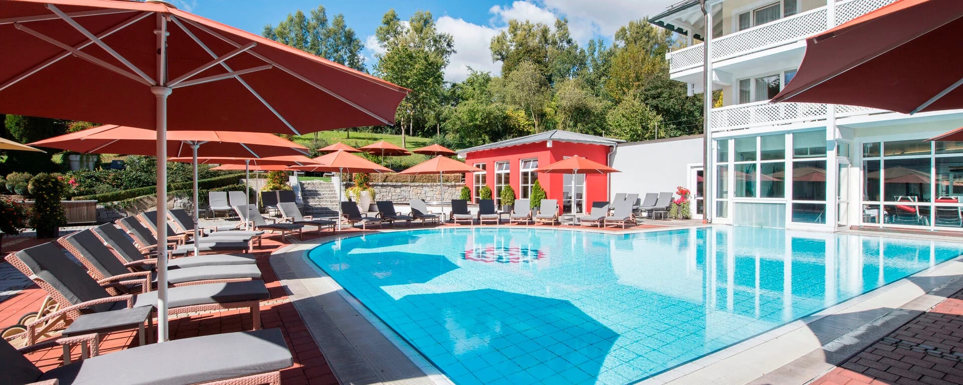 Außenpool mit Liegestühlen und Sonnenschirmen vor einem roten Gebäude und begrünter Böschung