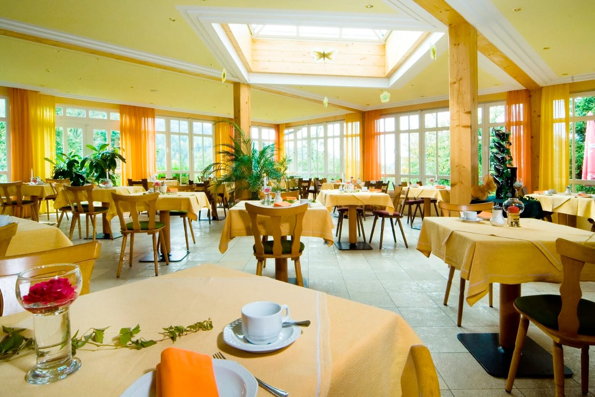 Heller Wintergarten mit gelben Vorhängen, Dachfenster, Grünpflanzen und gedeckten Tischen