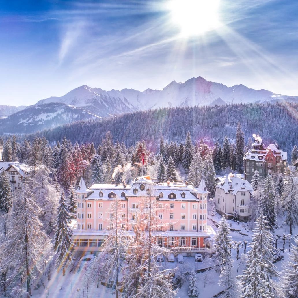 Verschneites Hotelgebäude zwischen schneebedeckten Tannen, dahinter Bergkette und tiefstehende Sonne