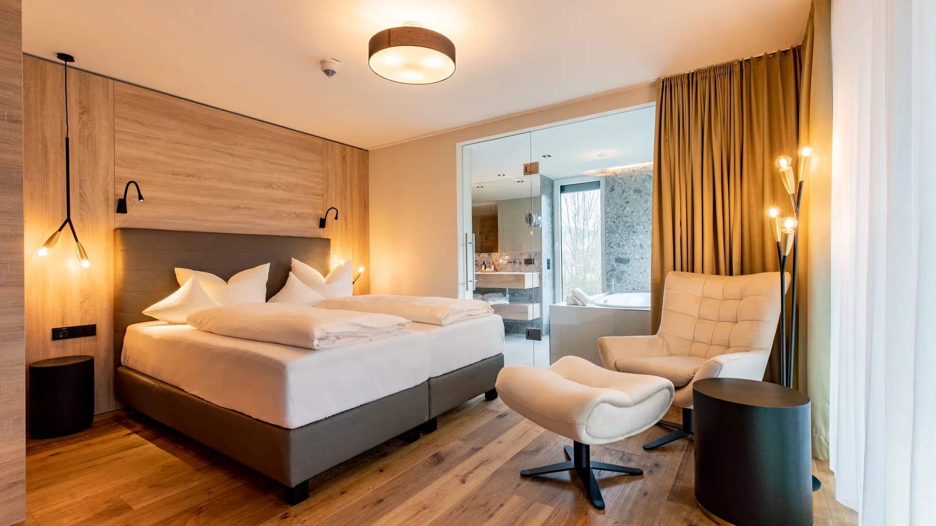 Modernes Zimmer mit Doppelbett, Kamin, Sessel und offenem Bad hinter Glaswand