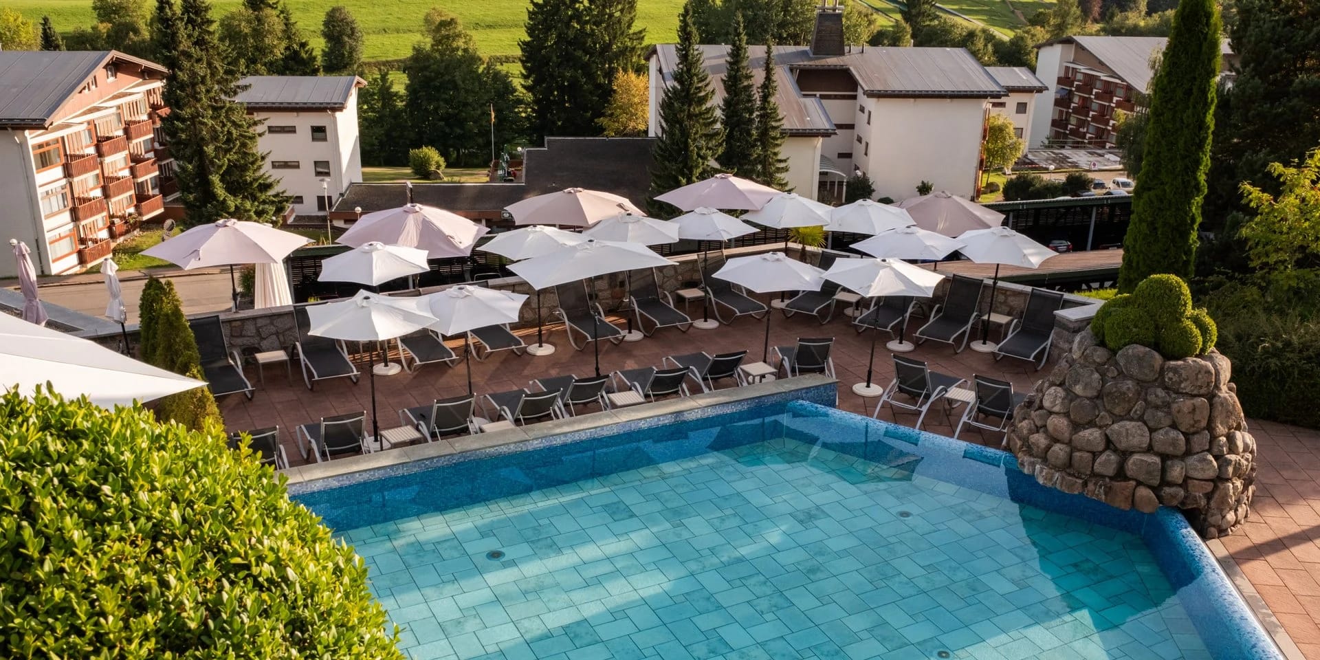 Blick von oben auf den Außenpool mit Sonnenschirmen und Liegereihen, dahinter die Hotelgebäude