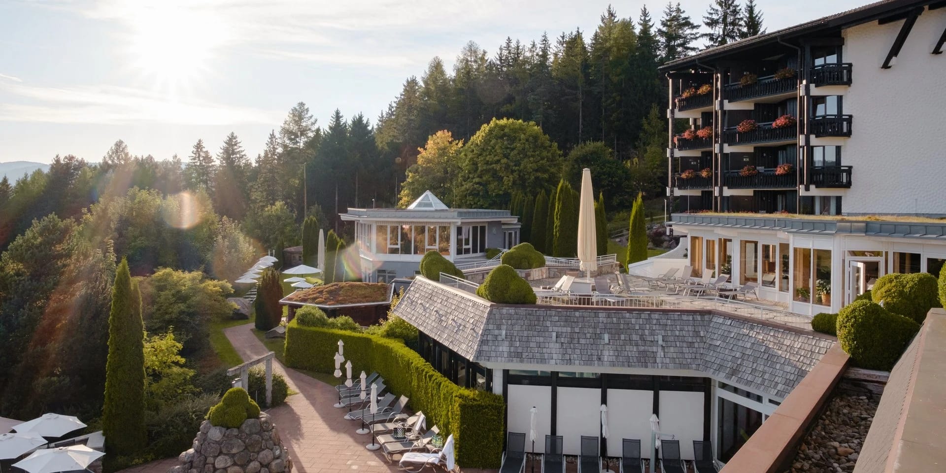 Blick über die terrassierte Gartenanlage auf den Hotelflügel mit dunklen Holzbalkonen, dahinter Wald
