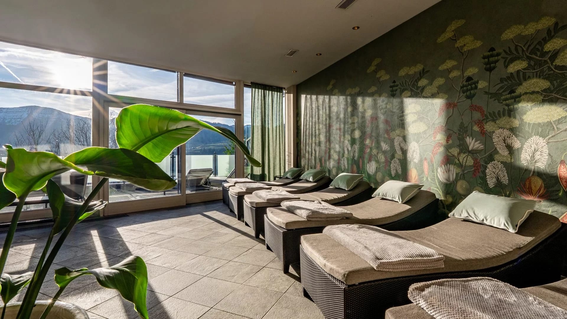 Ruheraum mit Liegen, Pflanzen und floraler Wandtapete, Fenster mit Bergblick im Sonnenlicht