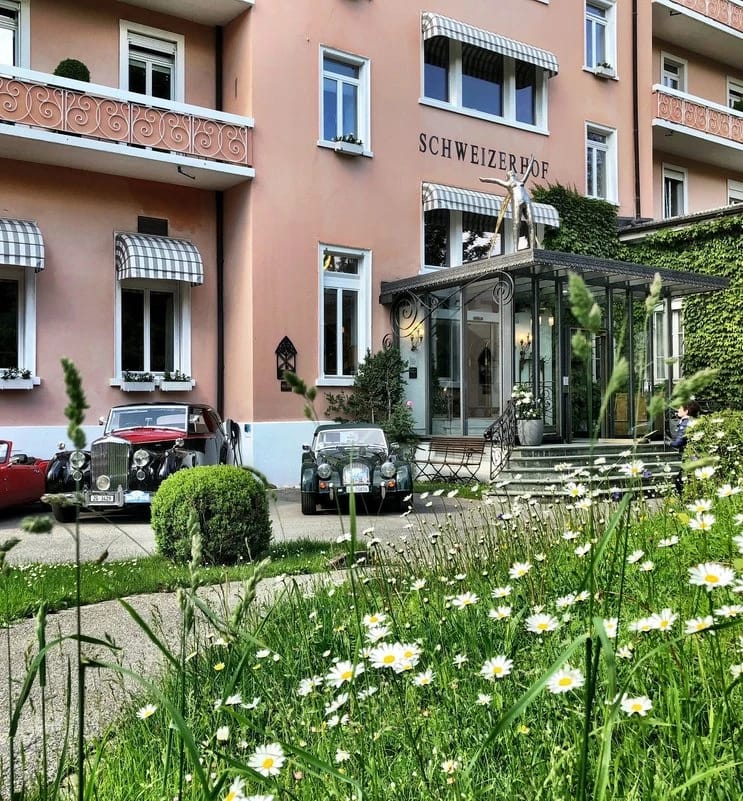 Rosafarbene Hausfassade mit Schriftzug Schweizerhof, gläsernem Eingangsvorbau und blühender Margeritenwiese davor