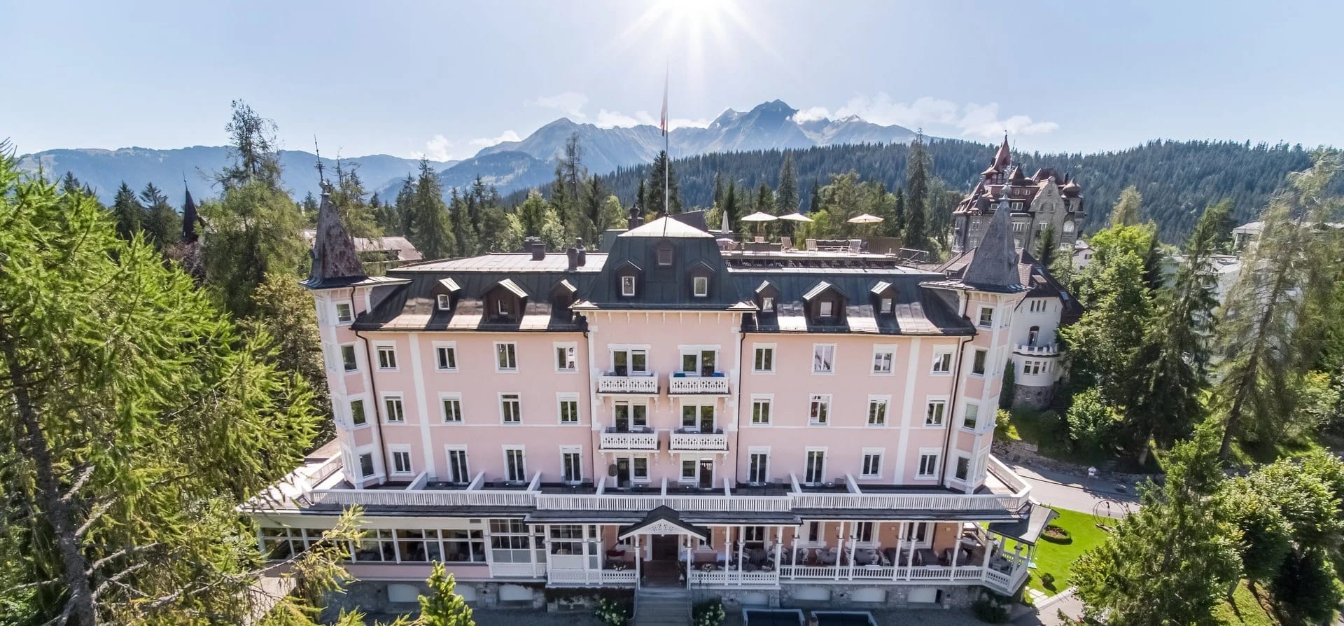 Luftaufnahme des Schweizerhof Flims mit Mansarddach und Türmchen, umgeben von Nadelwald, dahinter die Bergkette