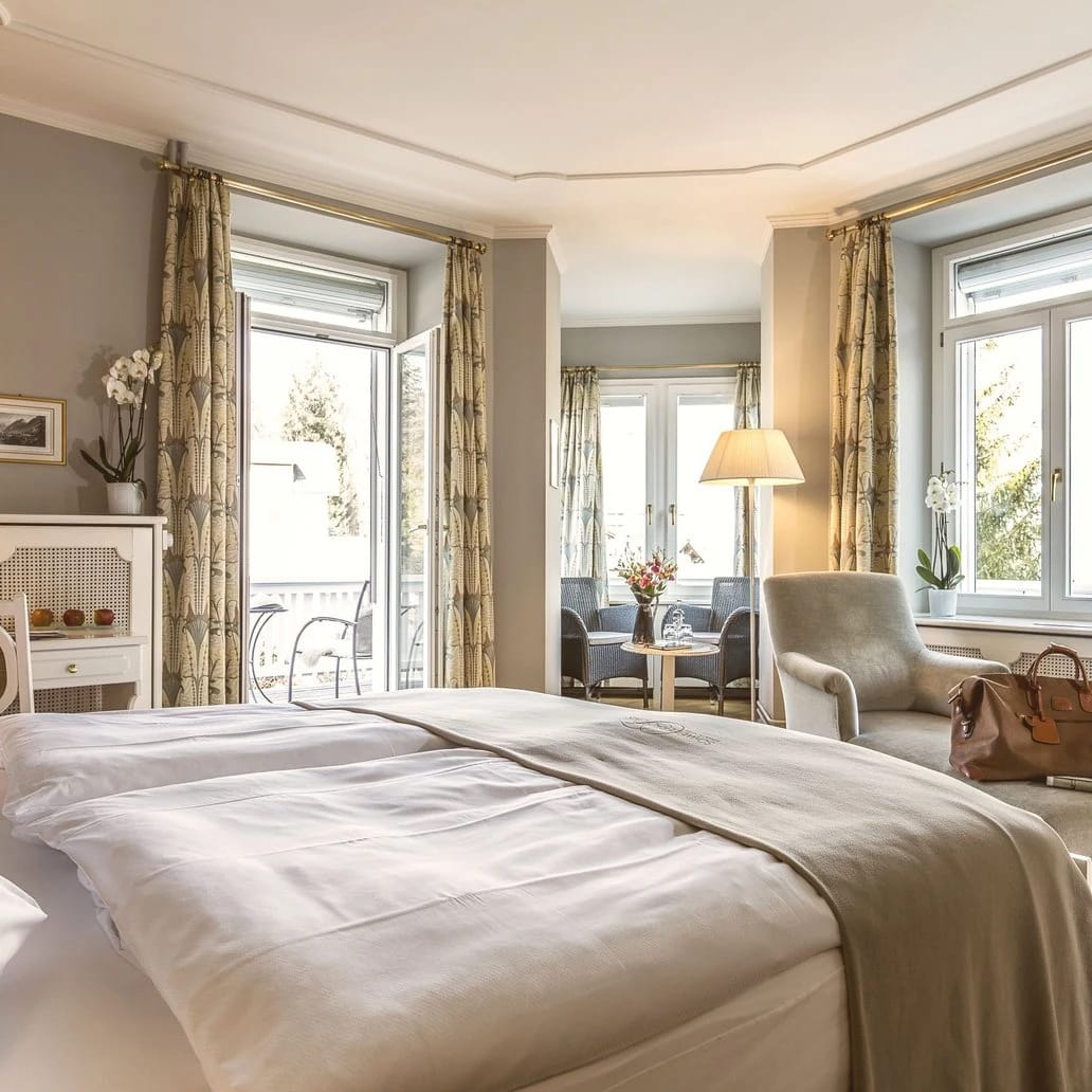 Doppelzimmer mit Doppelbett, zwei Fenstern mit gemusterten Vorhängen, Sessel und Tür zum kleinen Balkon