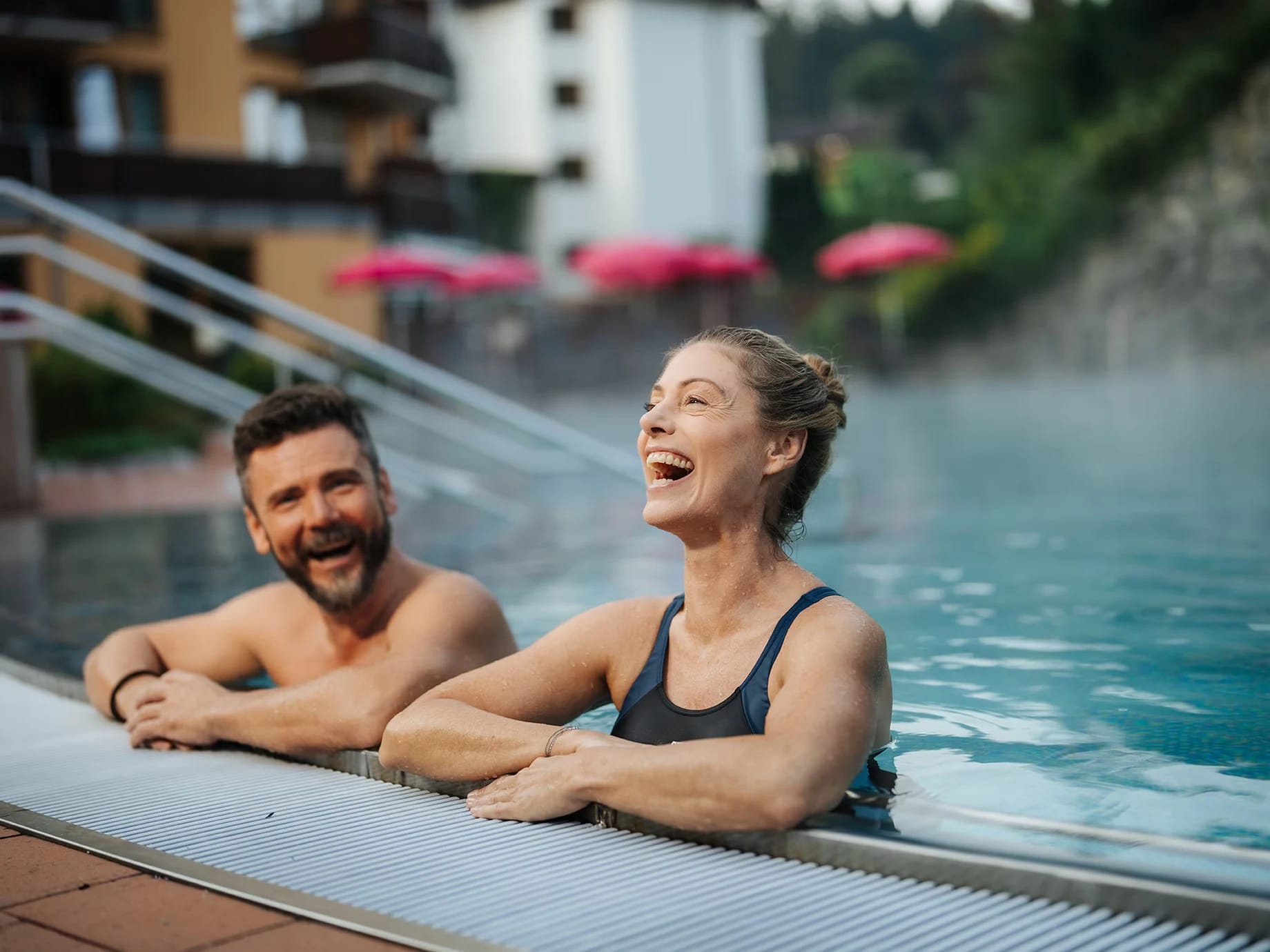 Paar am Rand des dampfenden Außenpools, dahinter das Hotelgebäude mit pinkfarbenen Sonnenschirmen