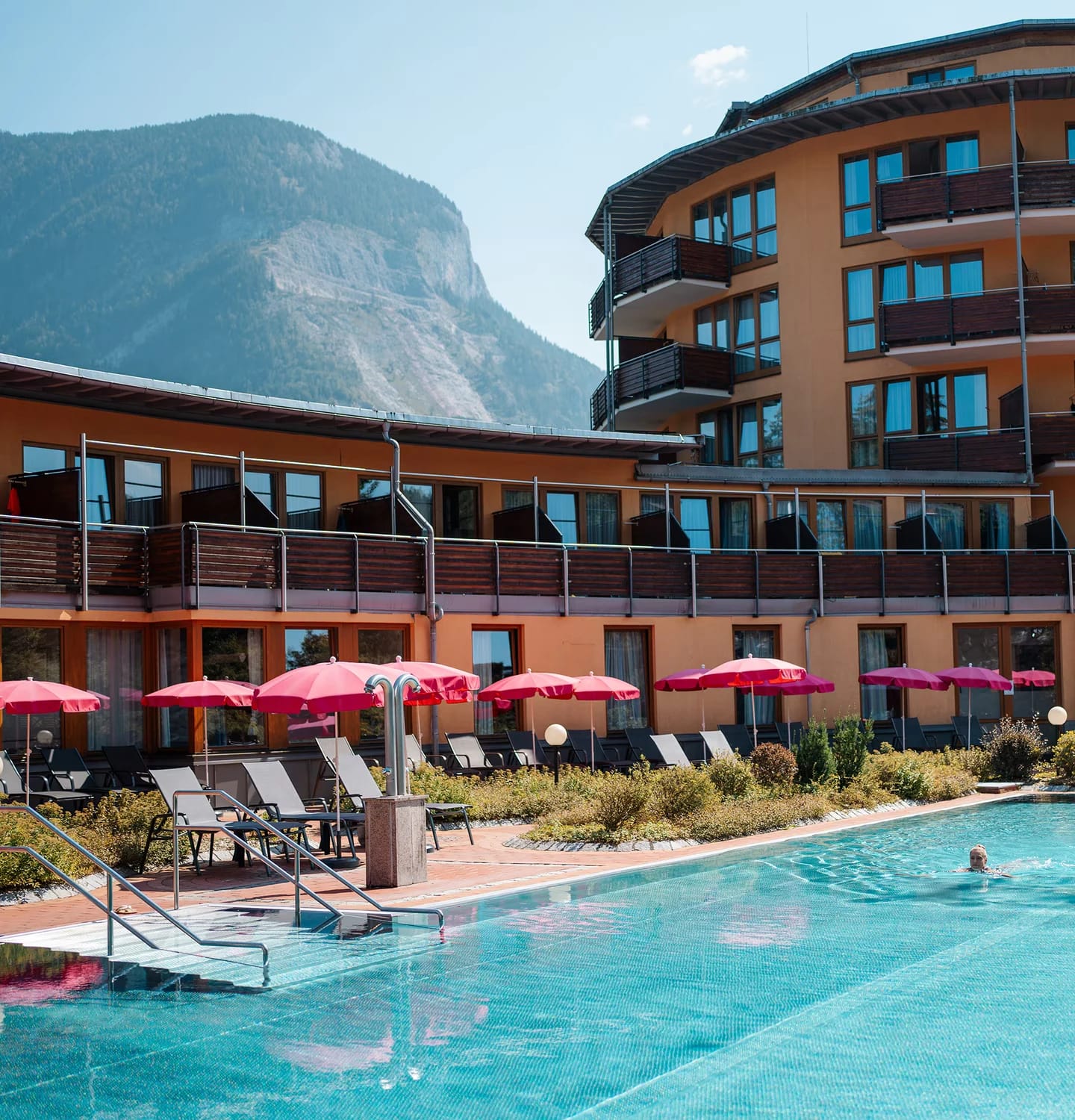 Außenansicht des Hotels mit geschwungener Fassade und Balkonen, davor der Außenpool mit Liegestühlen und pinkfarbenen Sonnenschirmen, dahinter ein Berg