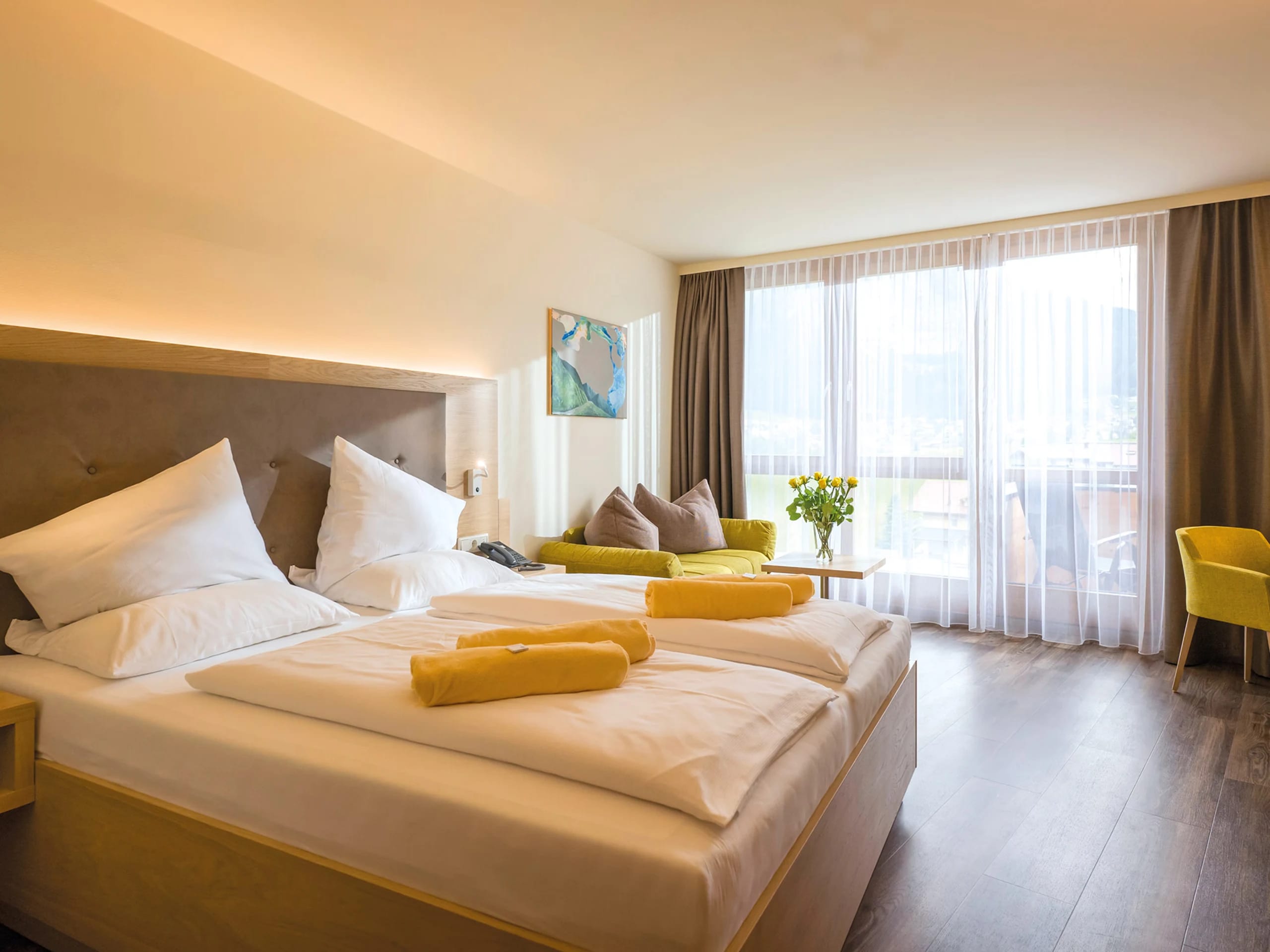 Doppelzimmer mit breitem Bett, Sitzecke, gelben Akzenten und bodentiefer Fenstertür zum Balkon