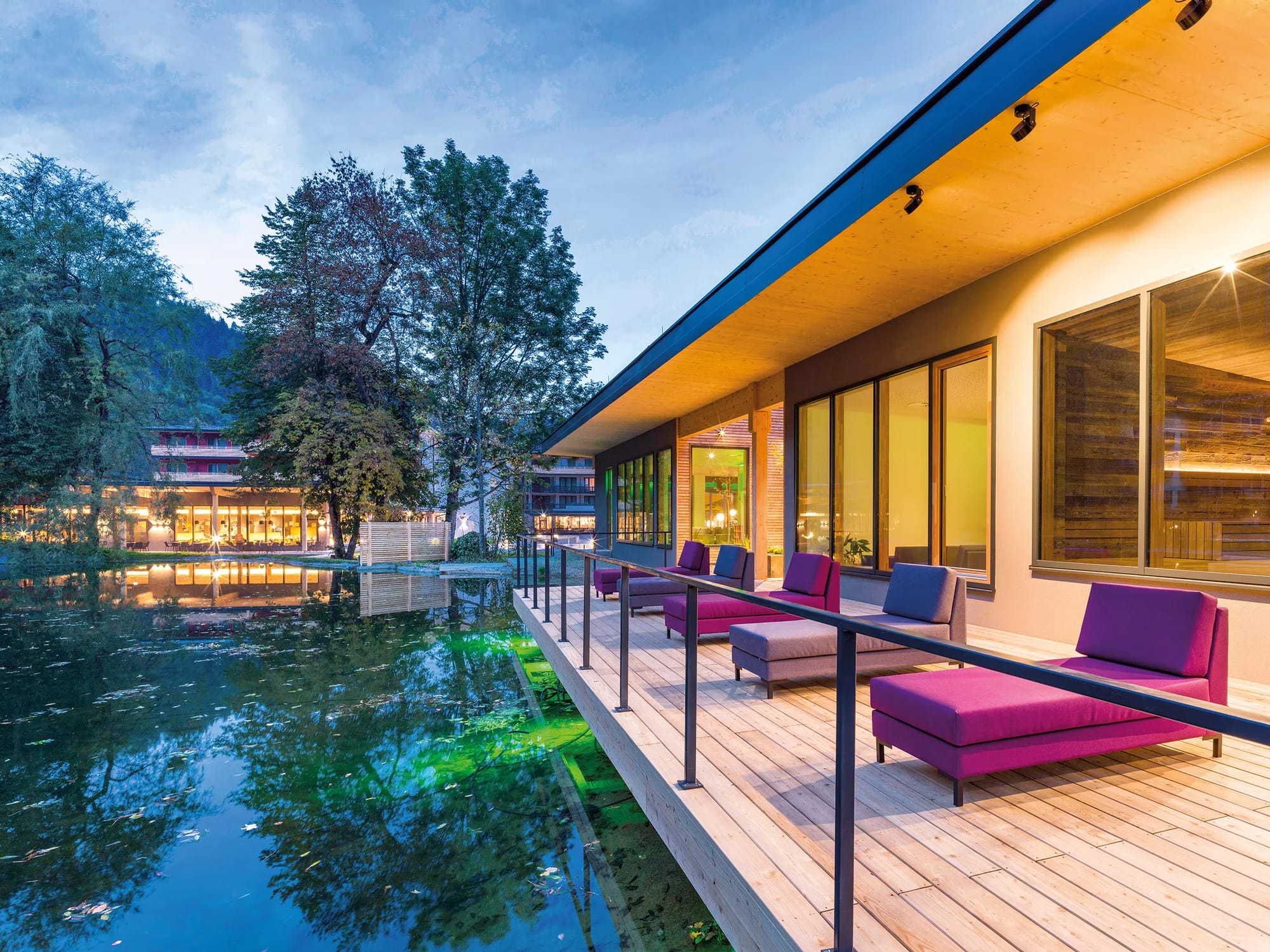 Holzterrasse mit Liegen am Teich in der Abenddämmerung, beleuchtetes Hotelgebäude im Hintergrund