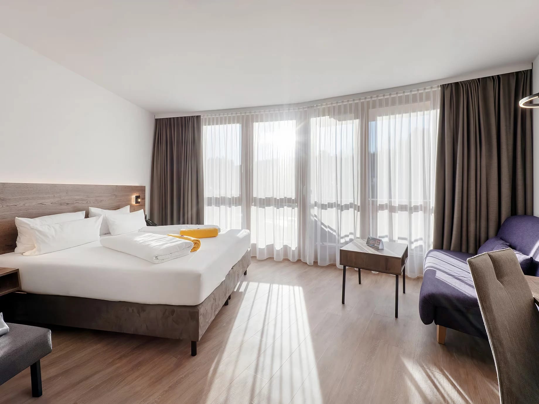 Helles Doppelzimmer mit großer Fensterfront, Bett, Sessel und Holzboden