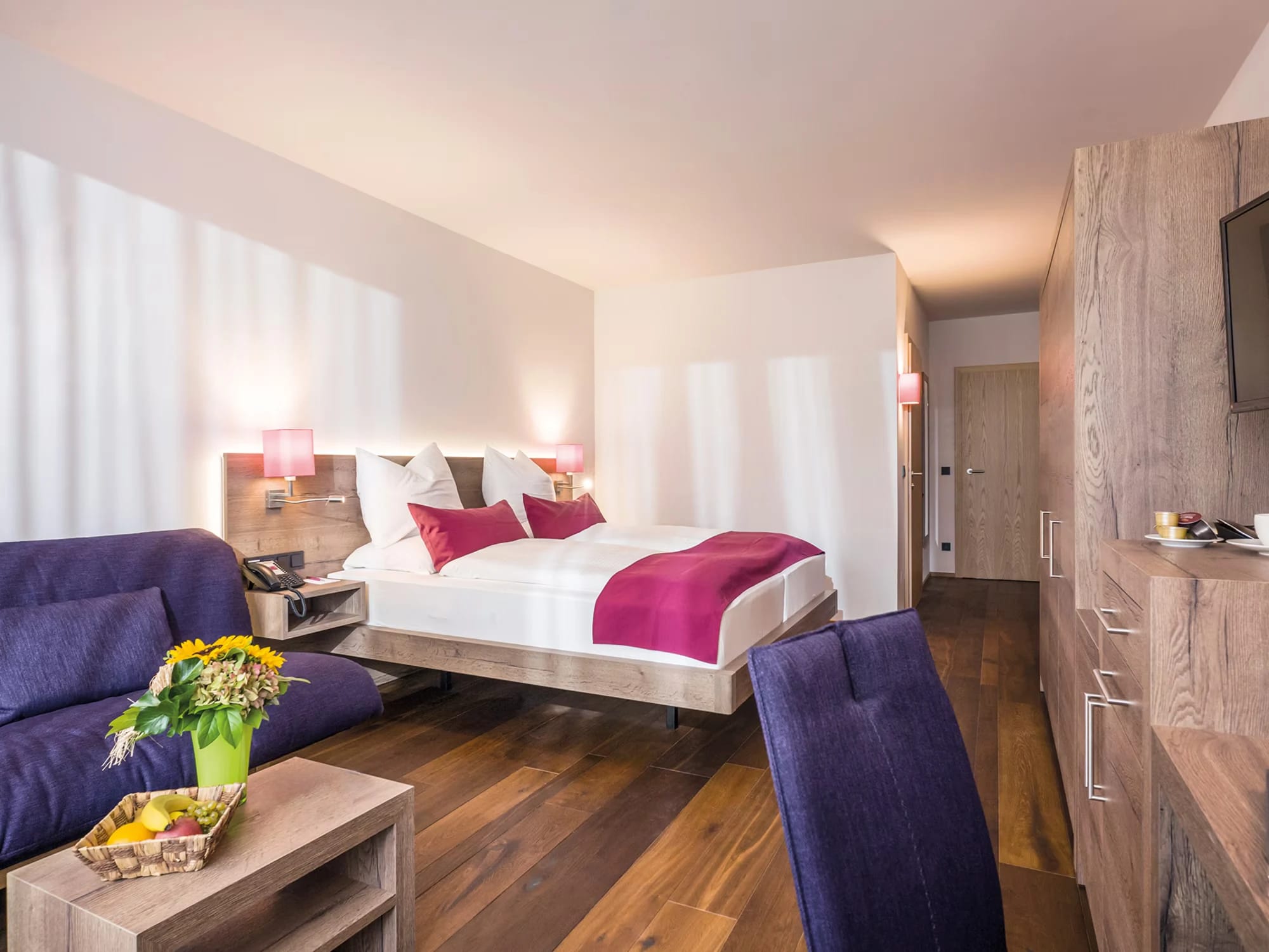 Helles Doppelzimmer mit Holzboden, roter Tagesdecke und großem Fenster