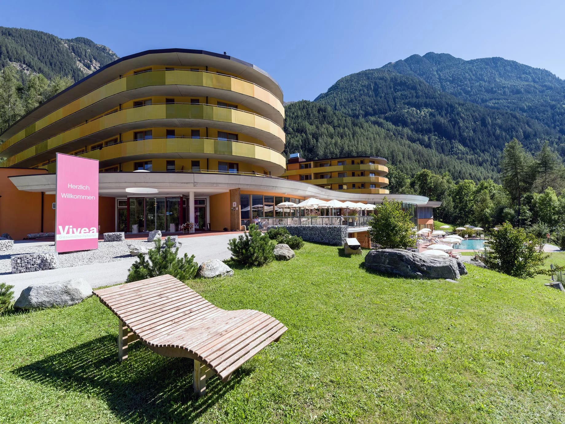 Außenansicht des Hotels mit geschwungener Fassade, Liegewiese und Bergen im Sommer