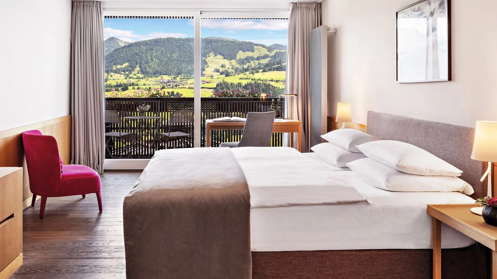 Doppelzimmer mit Doppelbett, pinkem Sessel und Balkon mit Blick auf grüne Berge