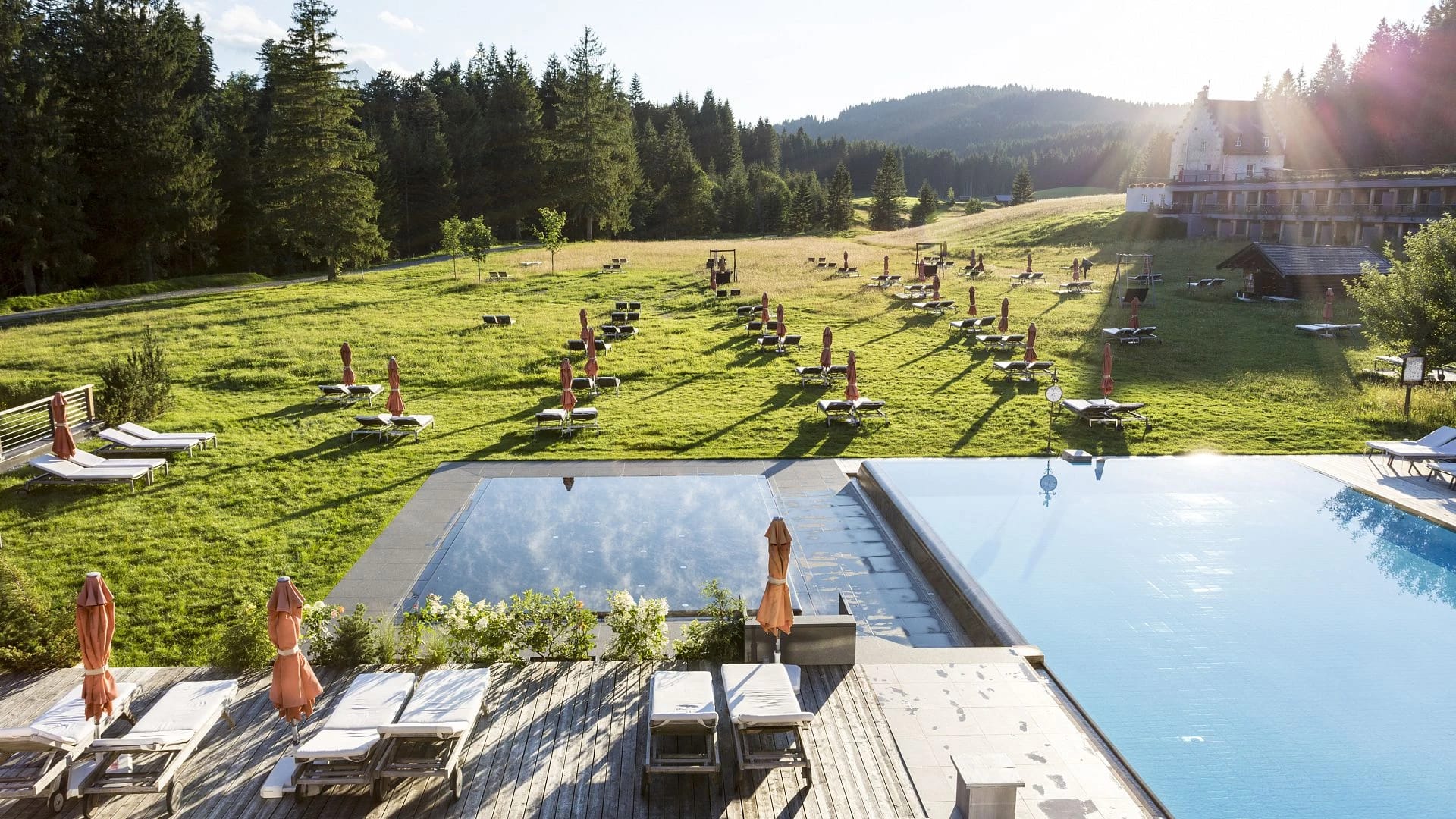 Zwei Außenpools mit Liegestühlen, dahinter eine große Liegewiese und ein Hotelflügel im Abendlicht