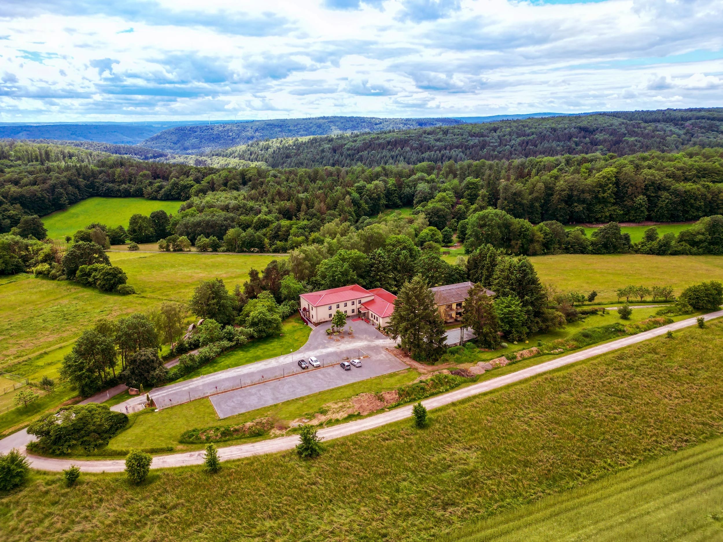 Luftaufnahme des Hauses mit Vorplatz und Zufahrt, ringsum Wiesen und bewaldete Hügel