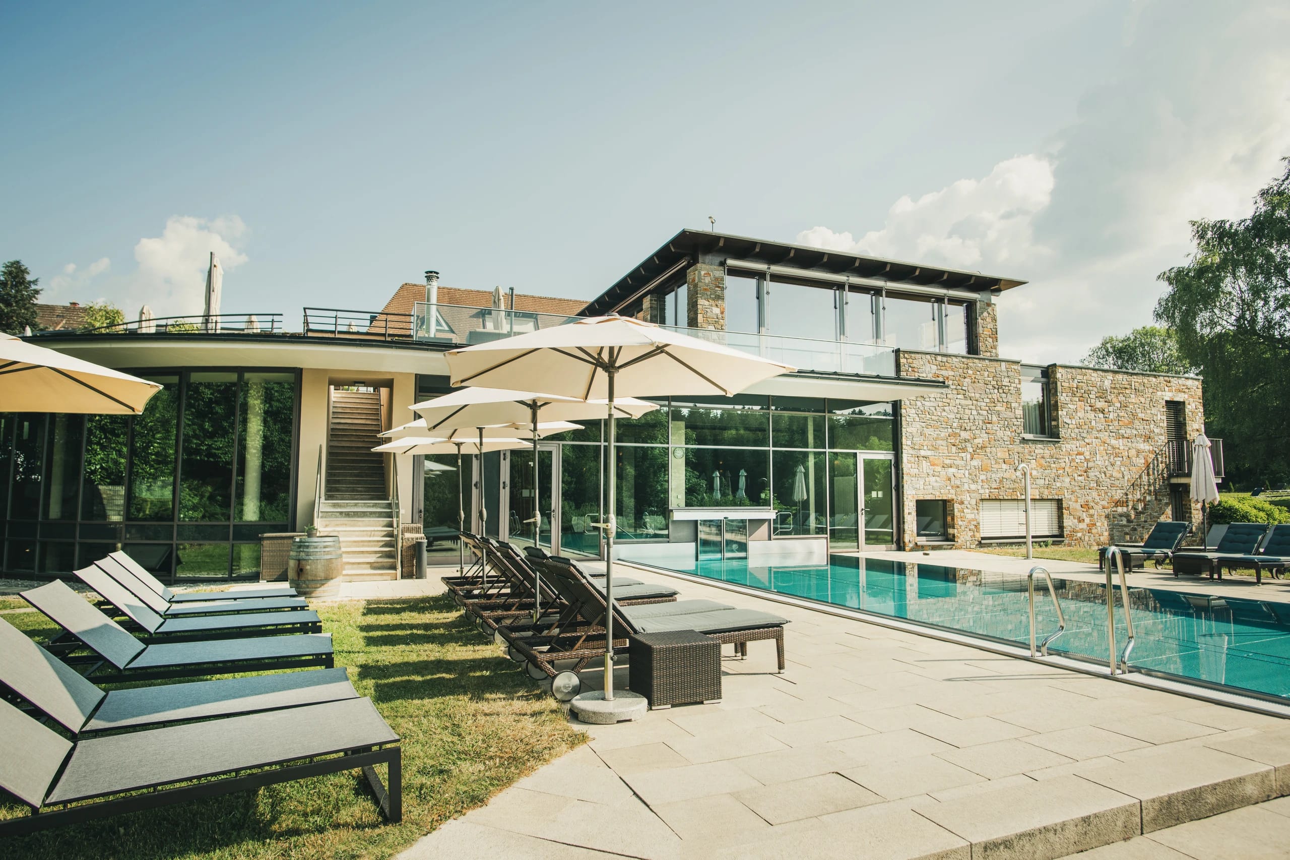 Außenansicht des Hotels aus Naturstein und Glas mit Außenpool, Sonnenschirmen und Liegen auf der Rasenfläche