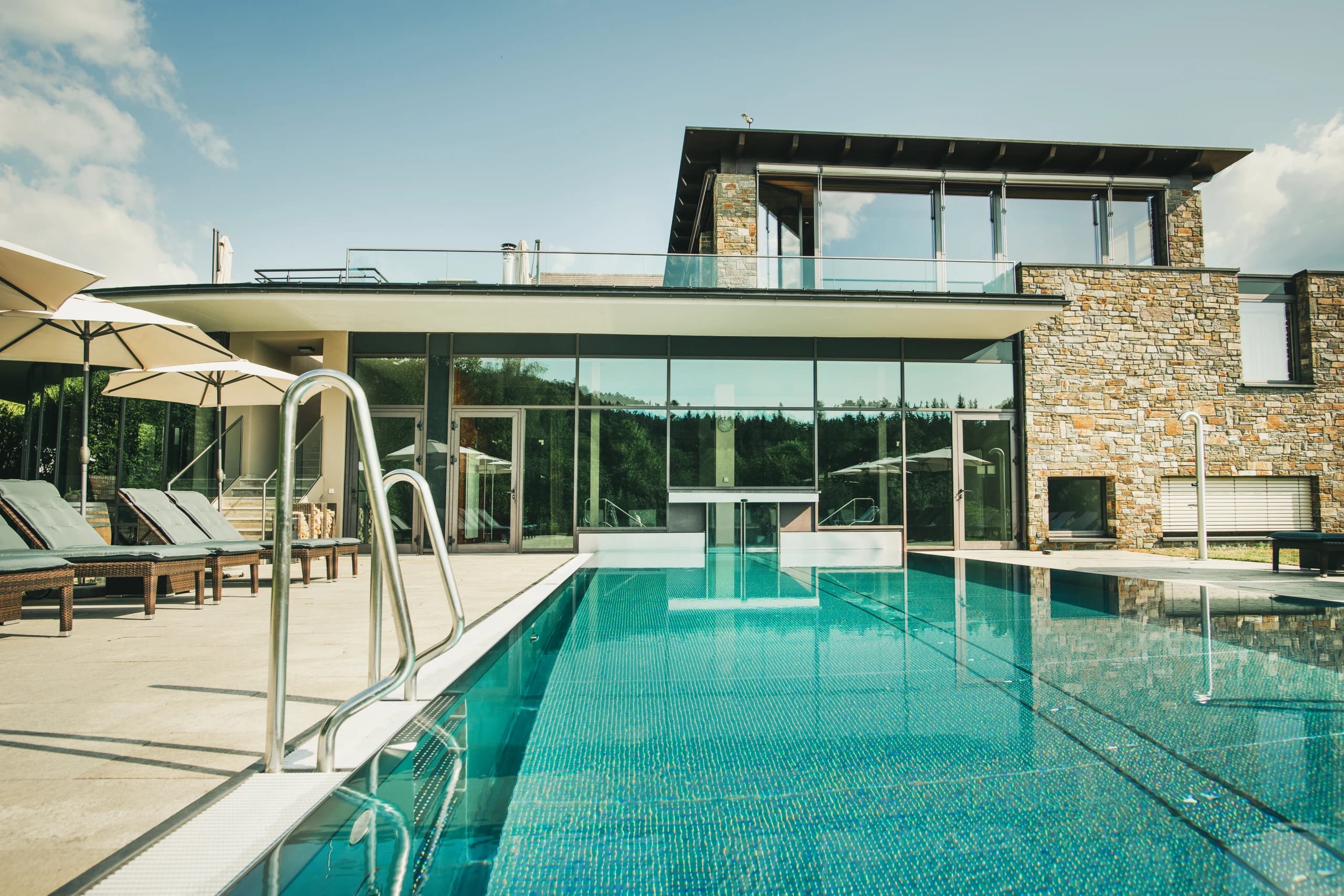Außenpool mit Sonnenschirmen und Liegestühlen vor der Glasfront des Hauses
