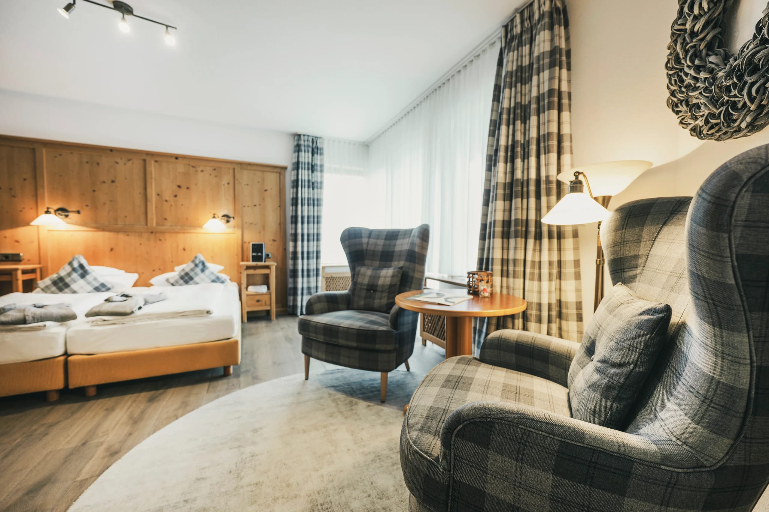 Hotelzimmer mit Doppelbett vor Holzkopfteil, zwei karierten Sesseln und rundem Beistelltisch