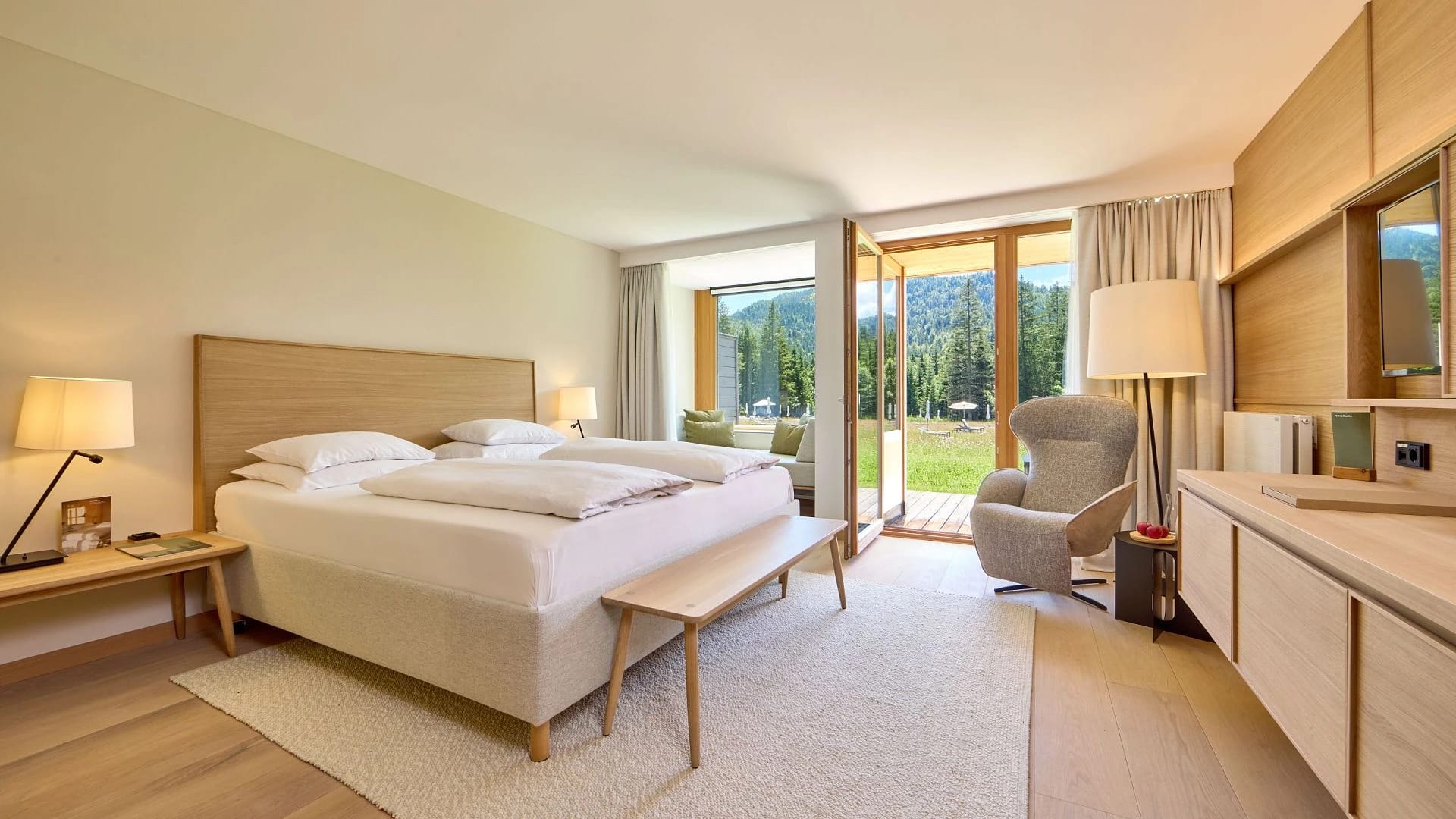 Helles Doppelzimmer mit Holzmöbeln, Sessel und Glastür zur Terrasse mit Blick ins Grüne