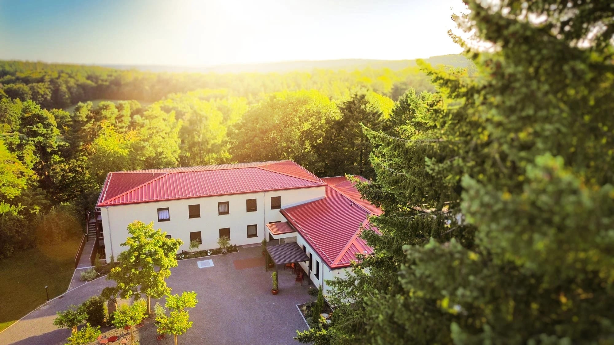 Luftaufnahme des Hauses mit rotem Dach, umgeben von Bäumen, im Hintergrund Wald