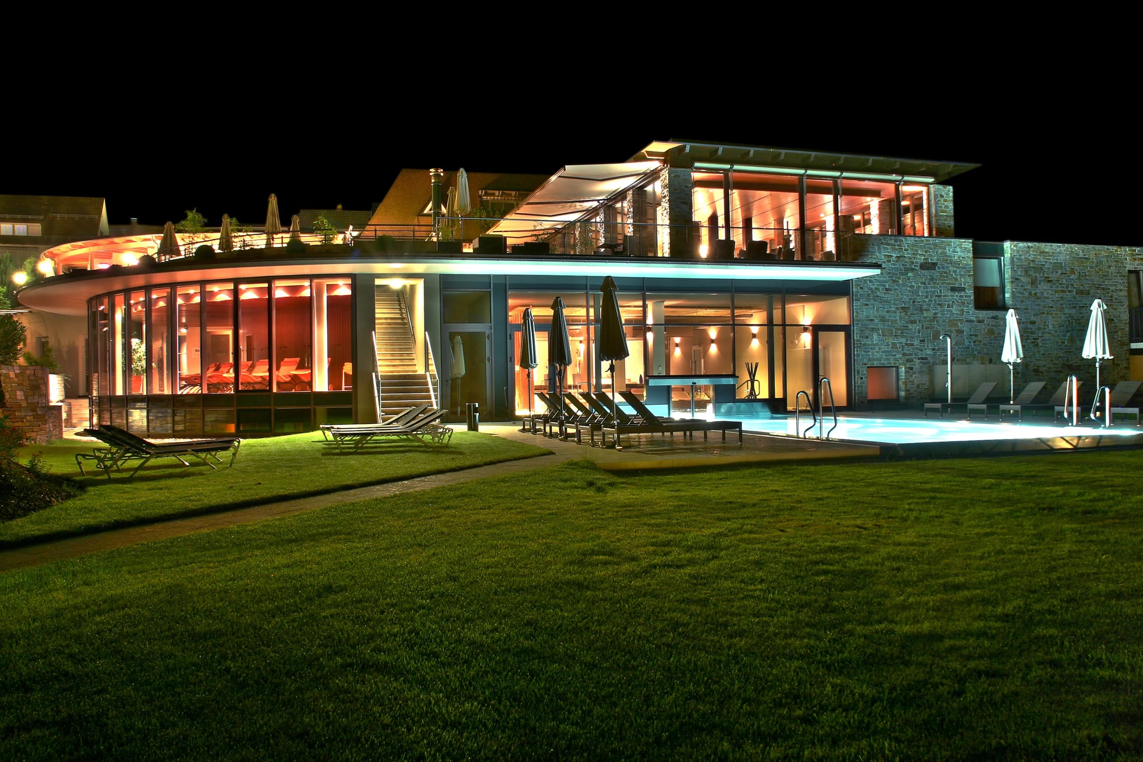Beleuchtete Hotelfassade am Abend mit Liegewiese, Terrasse und Pool