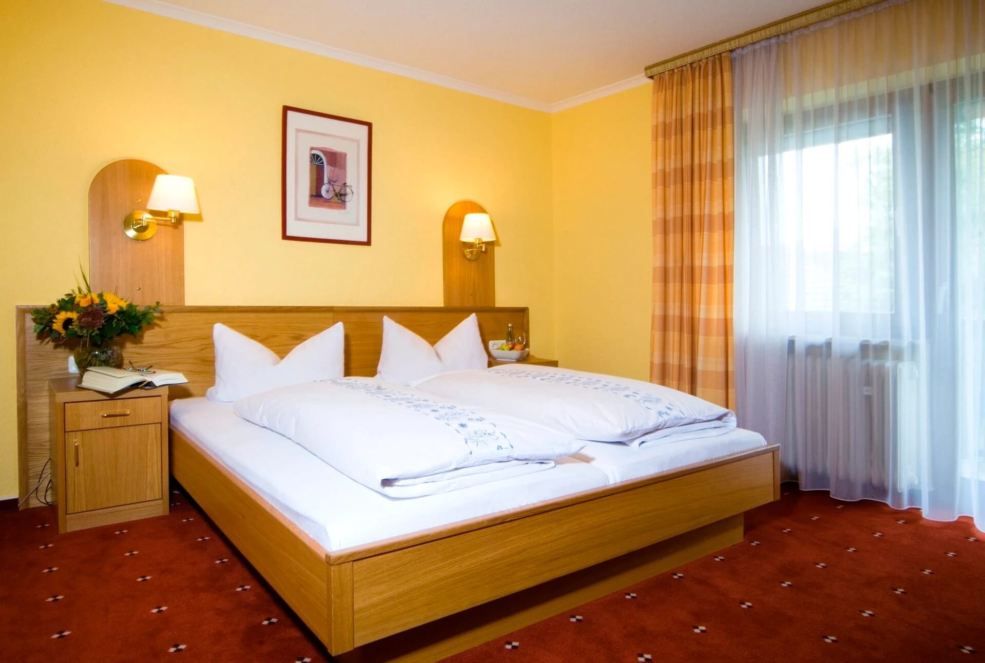 Doppelzimmer mit gelben Wänden, hellem Holzbett, weißer Bettwäsche und rotem Teppichboden
