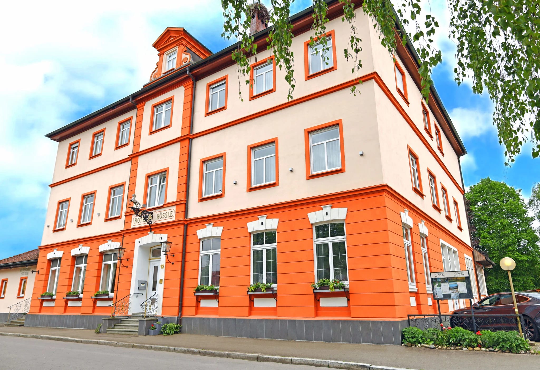 Orange gestrichenes Hotelgebäude mit weißen Fensterrahmen, Blumenkästen und Erker unter blauem Himmel