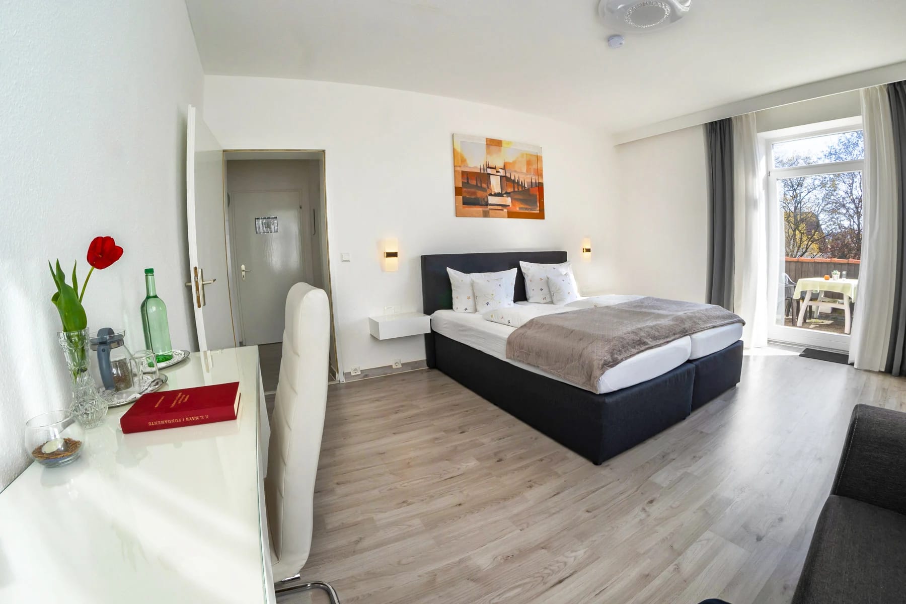 Helles Doppelzimmer mit Boxspringbett, weißem Schreibtisch und Tür zum Balkon