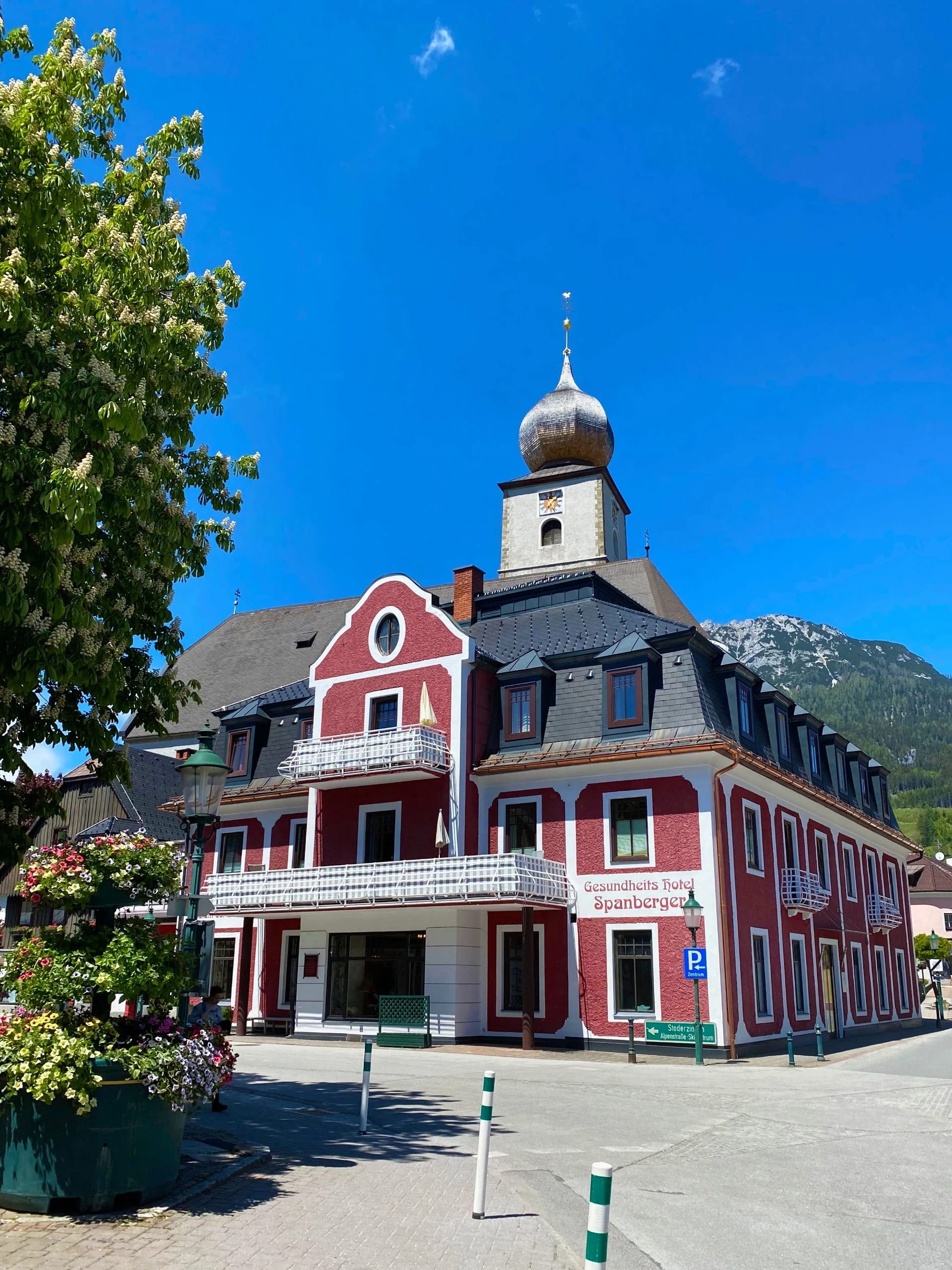 Rote Hausfassade mit Balkon und Hotelschild am Platz, daneben ein großer Laubbaum und der Kirchturm mit Zwiebelhaube