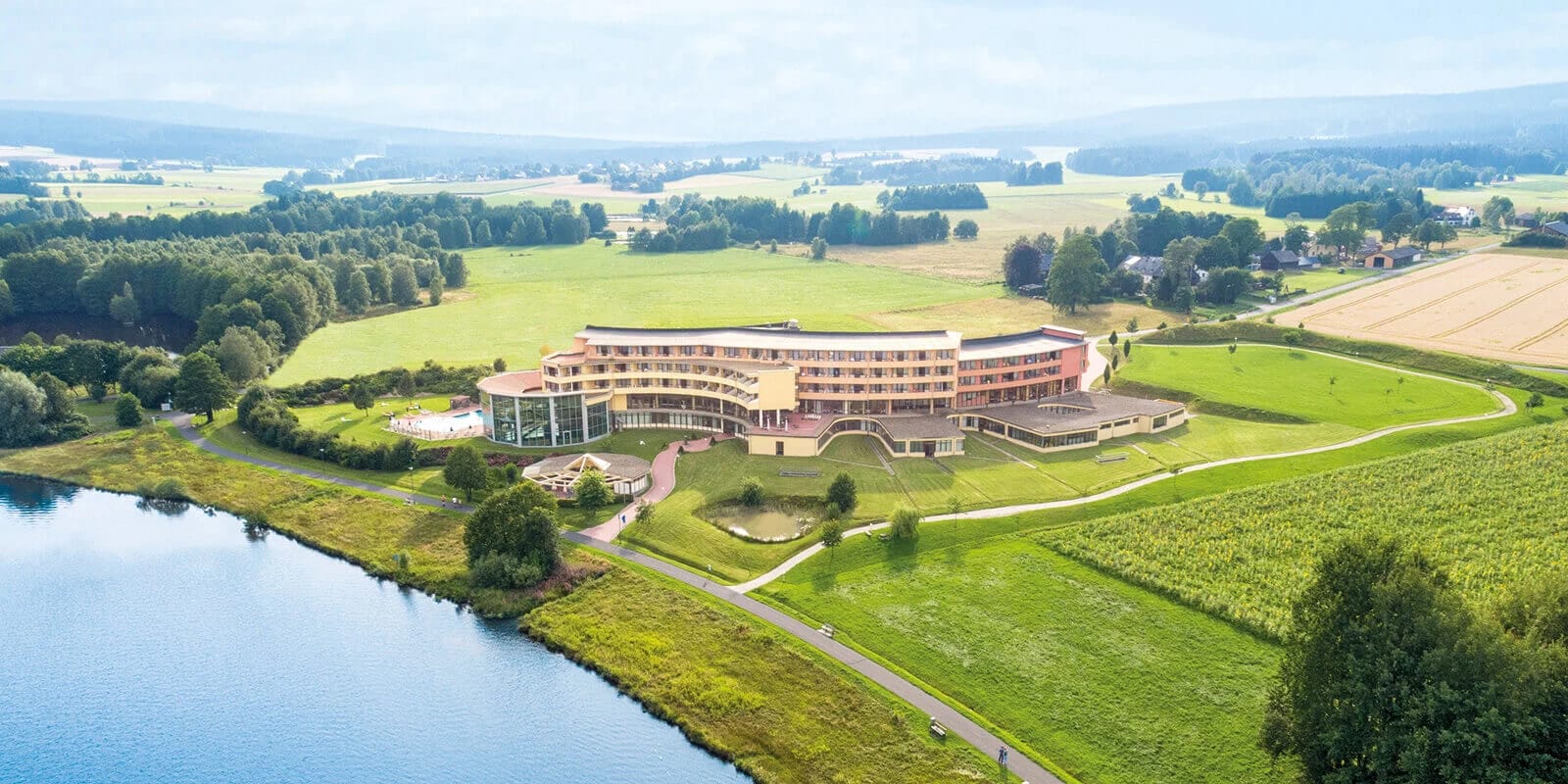 Luftaufnahme des Hotelgebäudes am Seeufer, umgeben von Wiesen und Wald