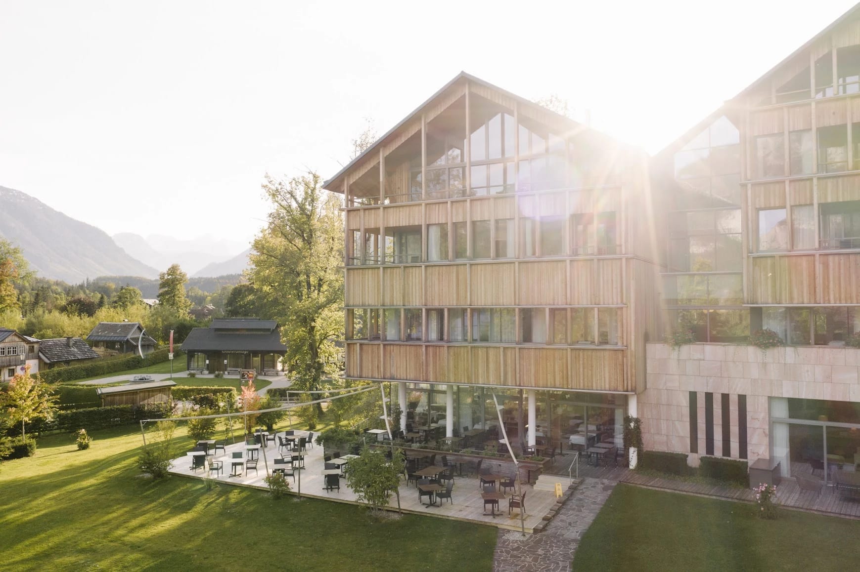 Hausansicht mit Holzfassade, Terrasse mit Tischen und Stühlen und Wiese im Abendlicht