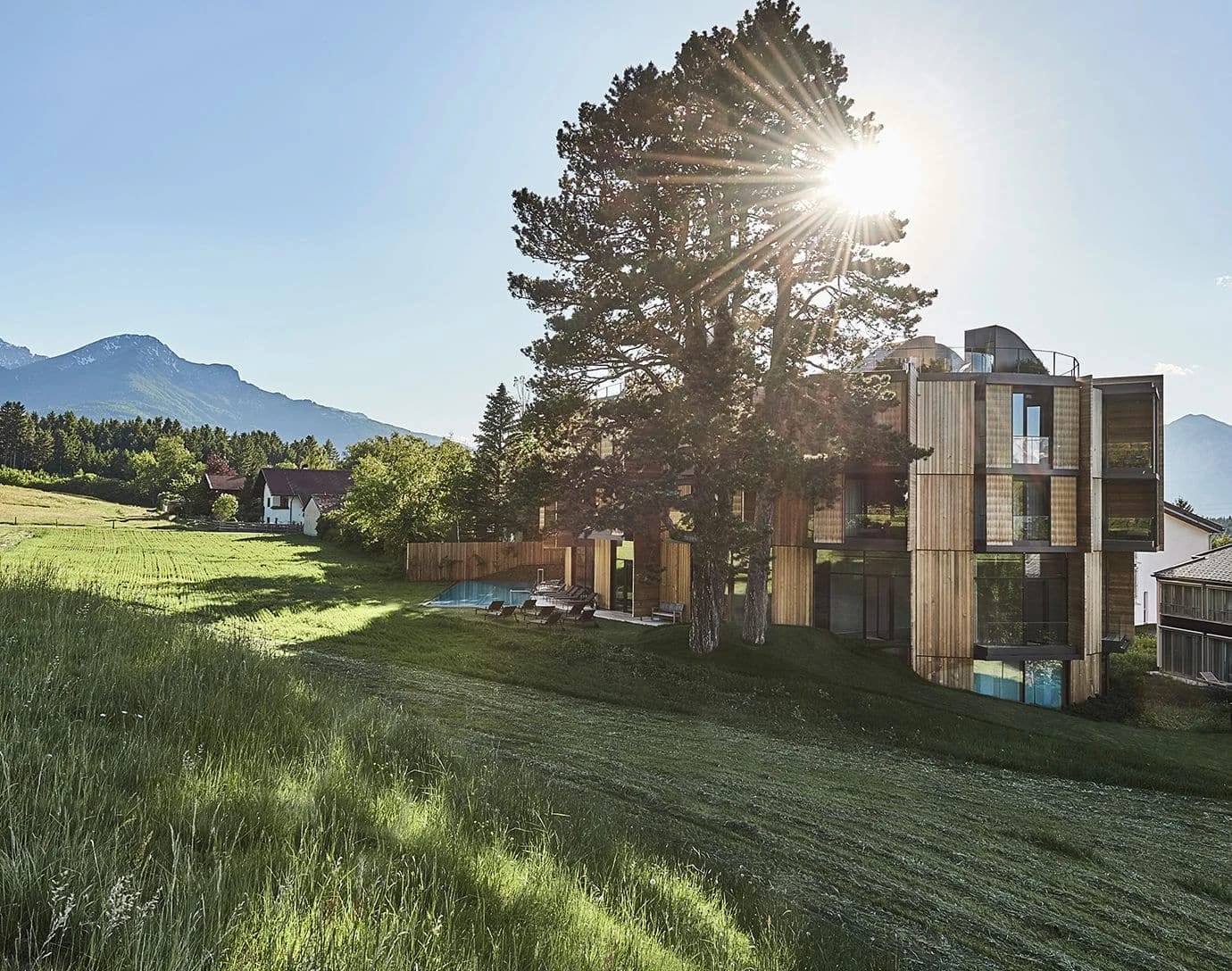 Holzfassade des Hauses mit Balkonen auf einer Wiese, daneben eine große Kiefer im Gegenlicht, im Hintergrund die Berge