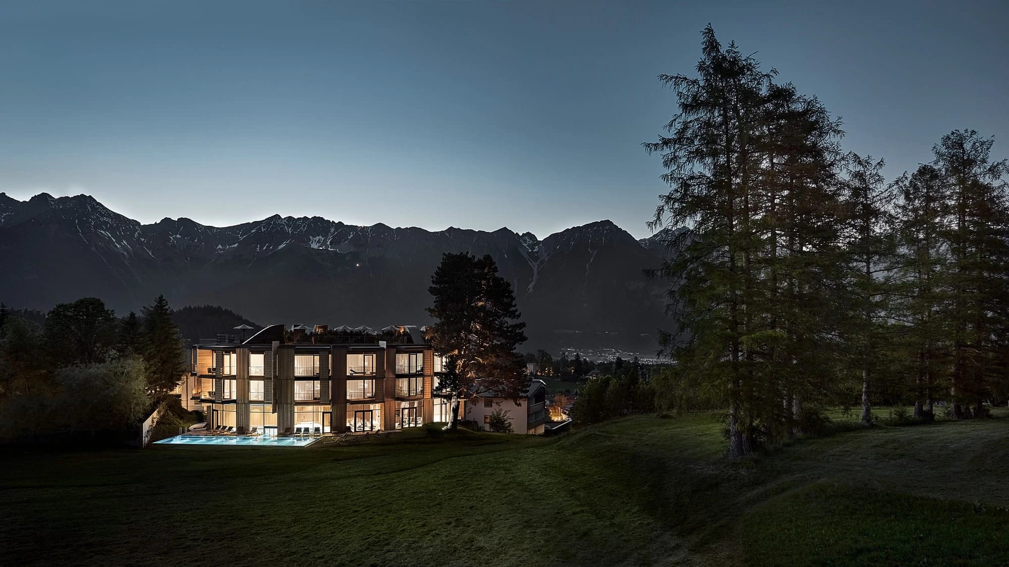 Beleuchtetes Haus in der Abenddämmerung, umgeben von Wiese und Bäumen, dahinter die Bergkette