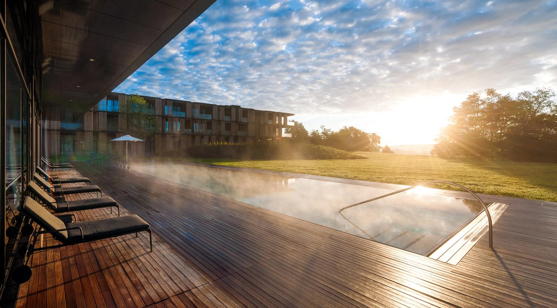 Holzterrasse mit Liegen am dampfenden Außenpool, dahinter der Gebäudeflügel im Morgenlicht