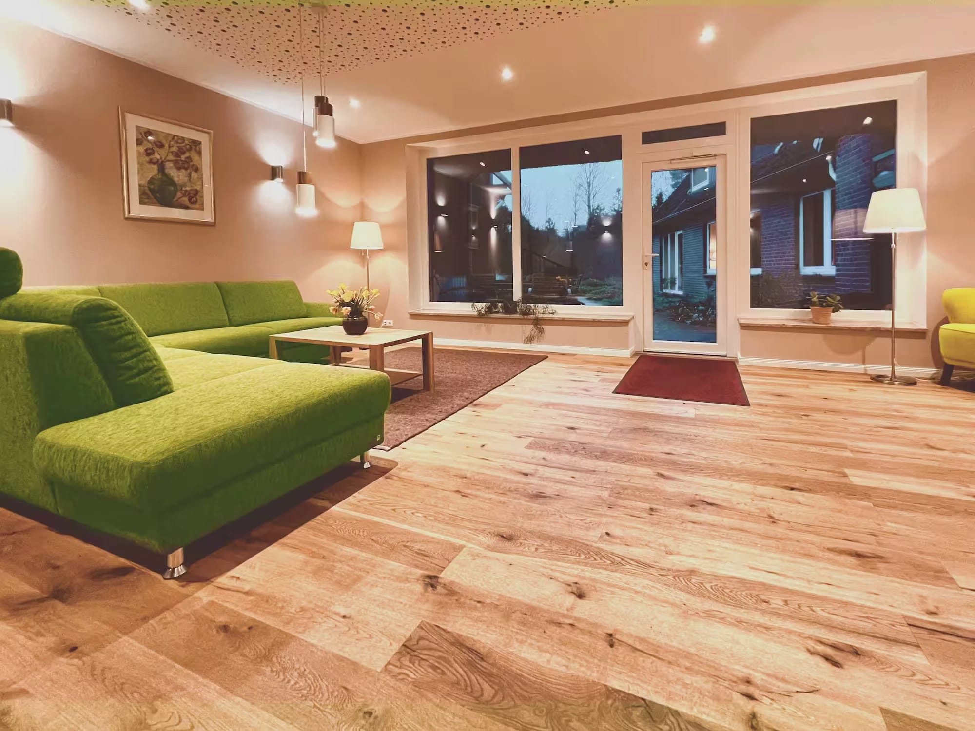 Lounge mit grüner Ecksofagarnitur, Holzdielen und Fensterfront am Abend