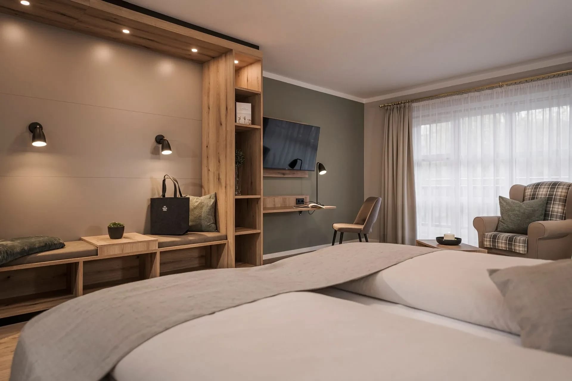 Zimmer mit Holzverkleidung hinter dem Bett, Fernseher, Schreibtisch und Sessel am Fenster