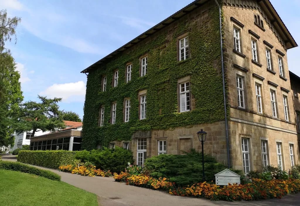 Efeubewachsenes Sandsteinhaus mit Blumenbeeten und Parkweg bei blauem Himmel