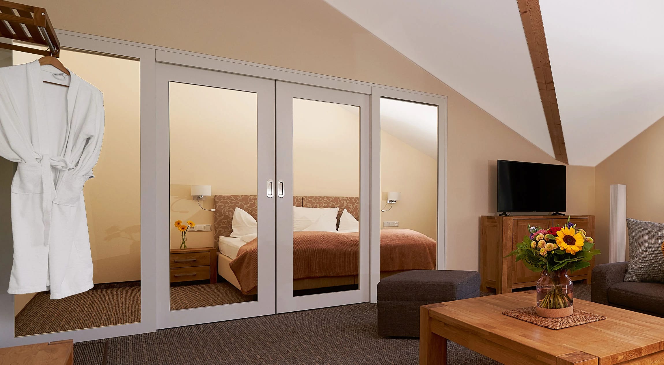 Suite mit Doppelbett hinter Spiegeltüren, Sitzecke, Bademantel und Blumen auf dem Holztisch