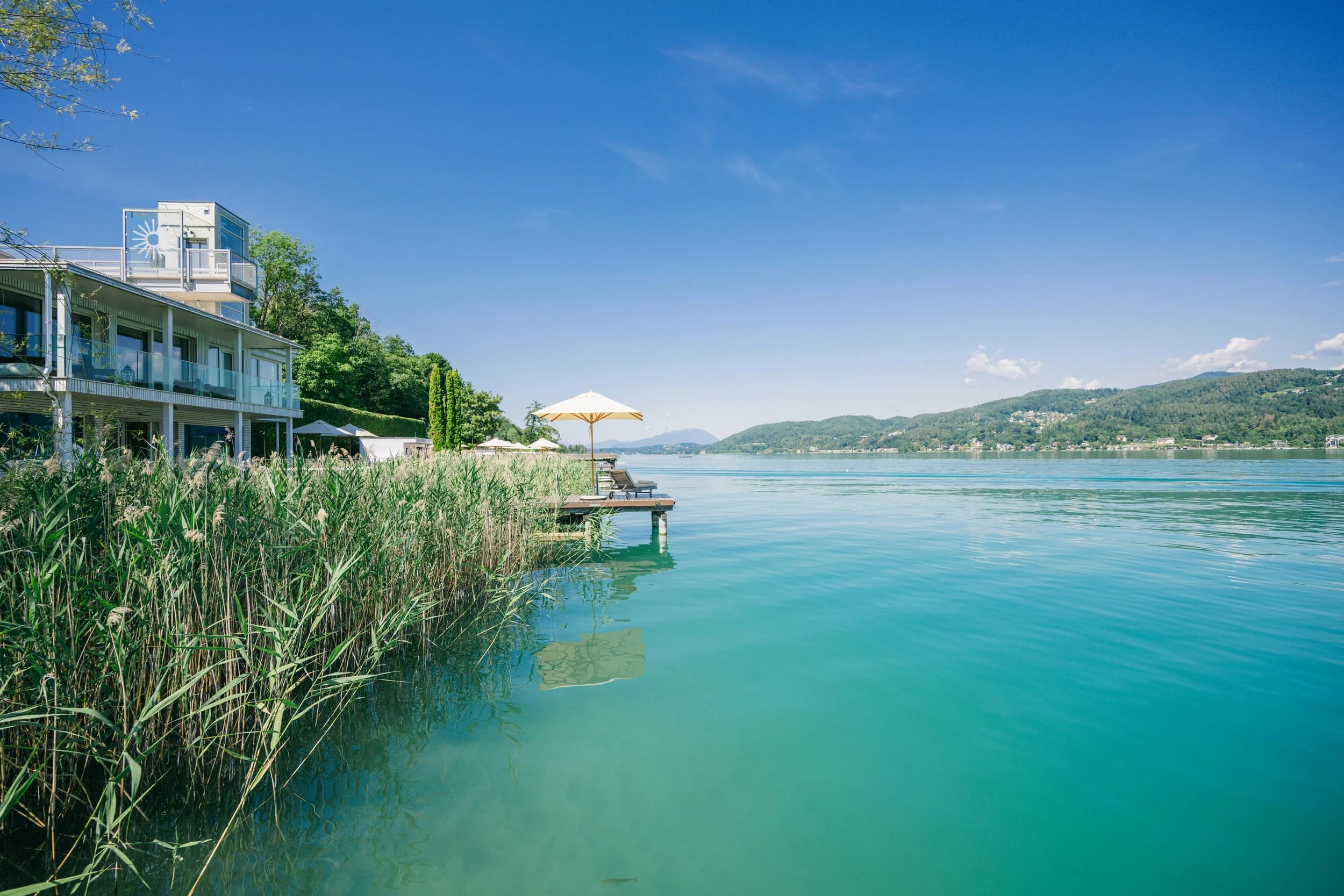 Blick vom See auf das Strandhaus mit Steg, Sonnenschirm und Schilfgürtel