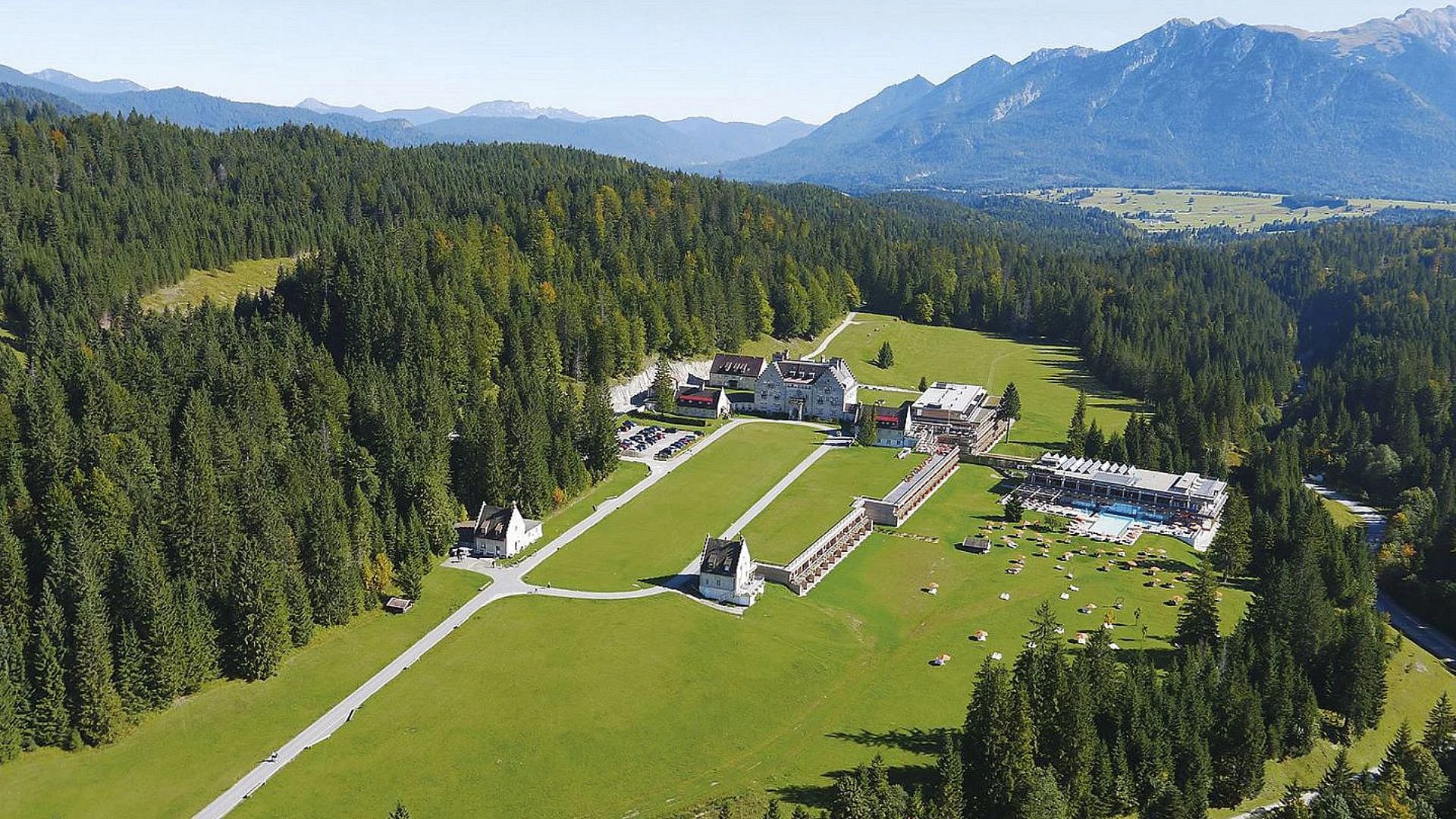 Luftaufnahme der Hotelanlage auf einer Waldlichtung mit Wiesen und Bergkette im Hintergrund