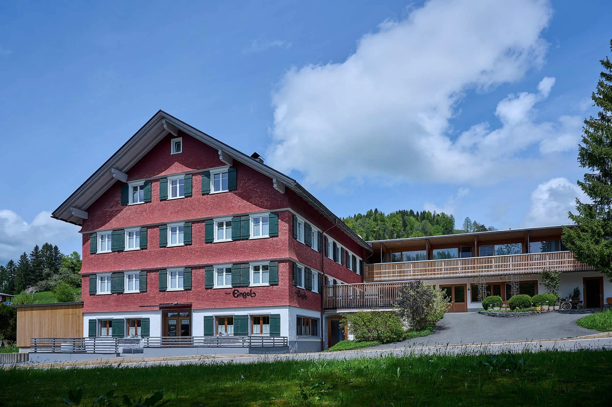 Rot gestrichenes Haupthaus mit grünen Fensterläden und Holzanbau, davor Wiese und Zufahrt im Sommer