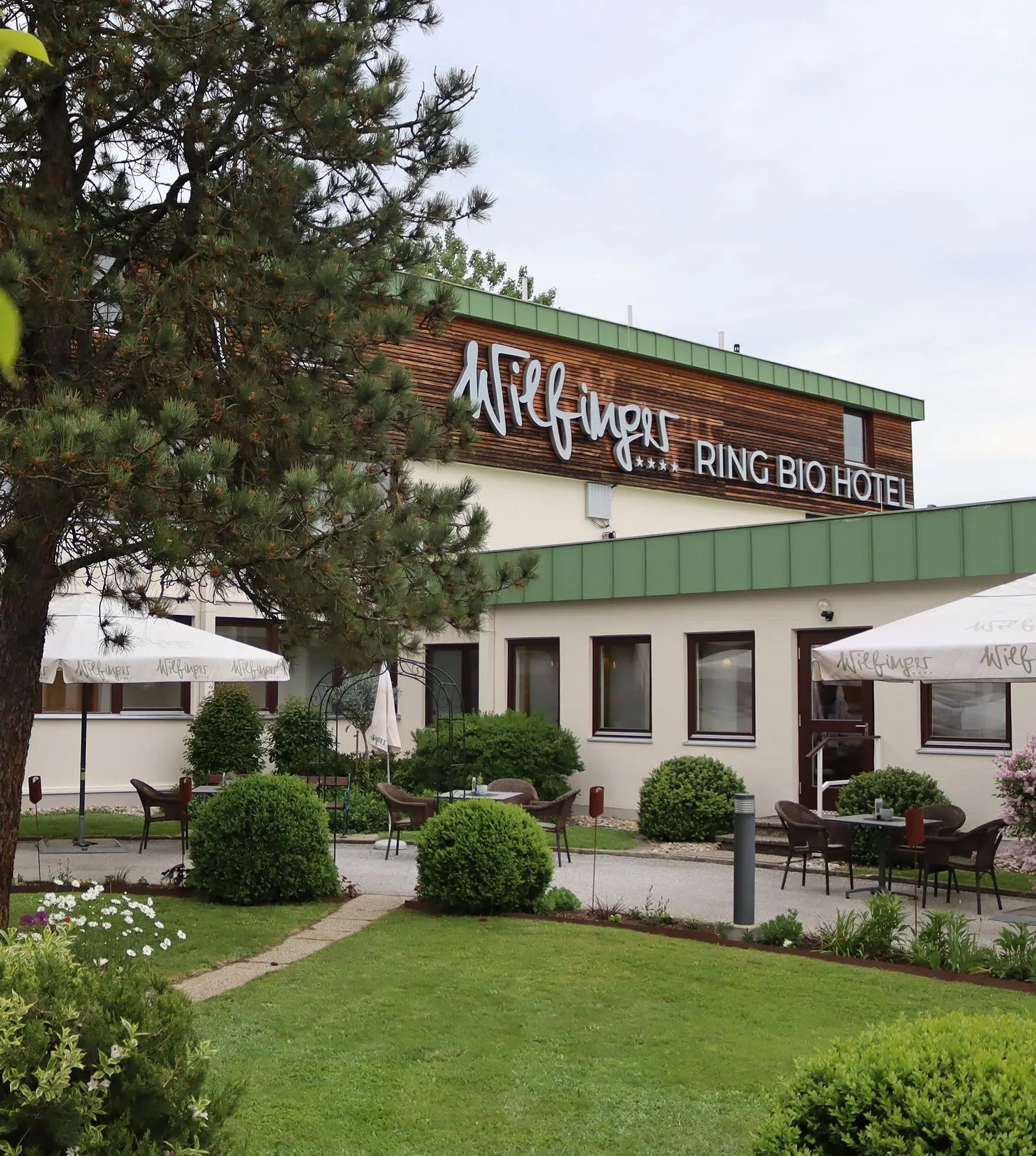 Eingangsbereich des Ring Bio Hotels mit Namensschild, Terrasse und Sonnenschirmen