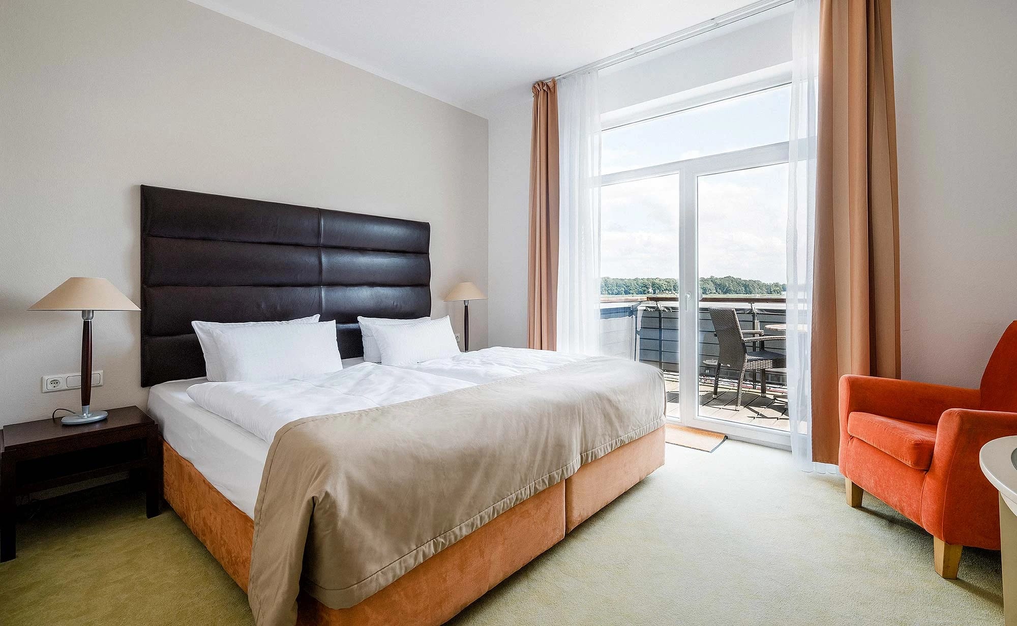Helles Doppelzimmer mit dunklem Kopfteil, orangefarbenem Sessel und Balkontür mit Blick auf den See
