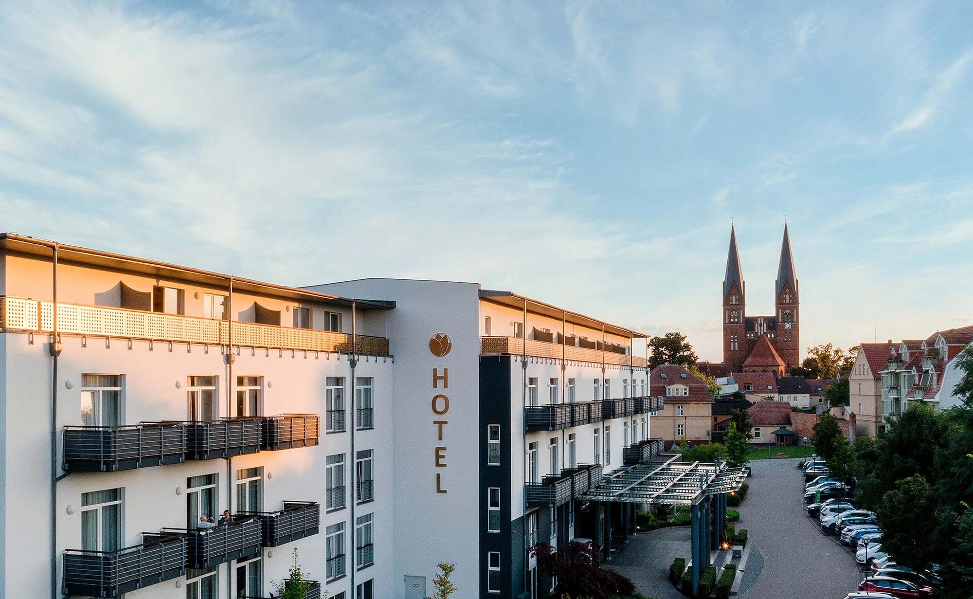 Hotelfassade mit Balkonen im Abendlicht, im Hintergrund die Doppeltürme einer Kirche