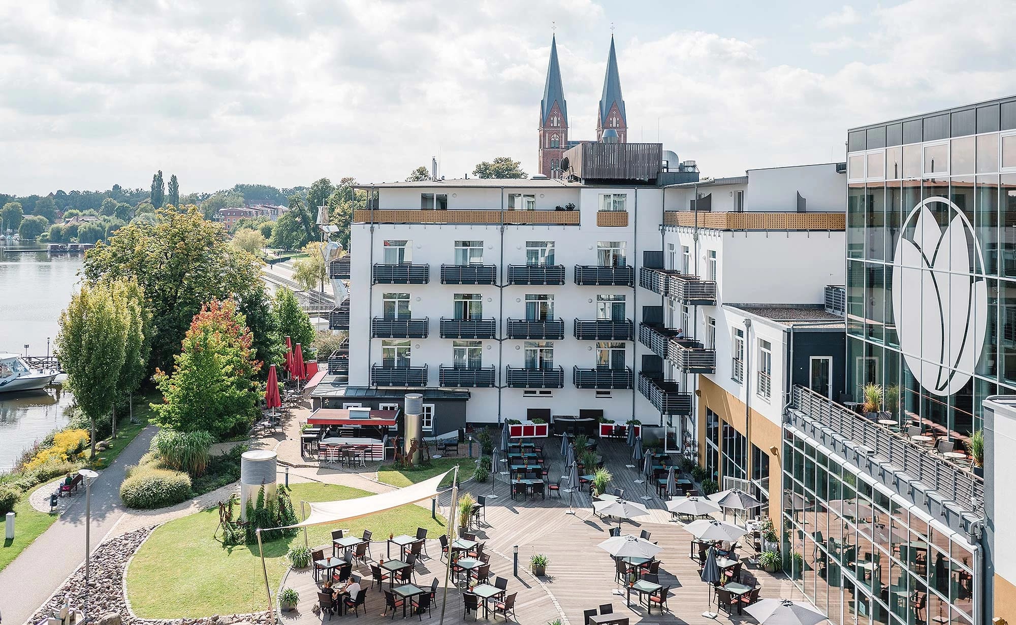 Terrasse mit Tischen und Sonnenschirmen zwischen den Hotelgebäuden, links der Ruppiner See, im Hintergrund zwei Kirchtürme