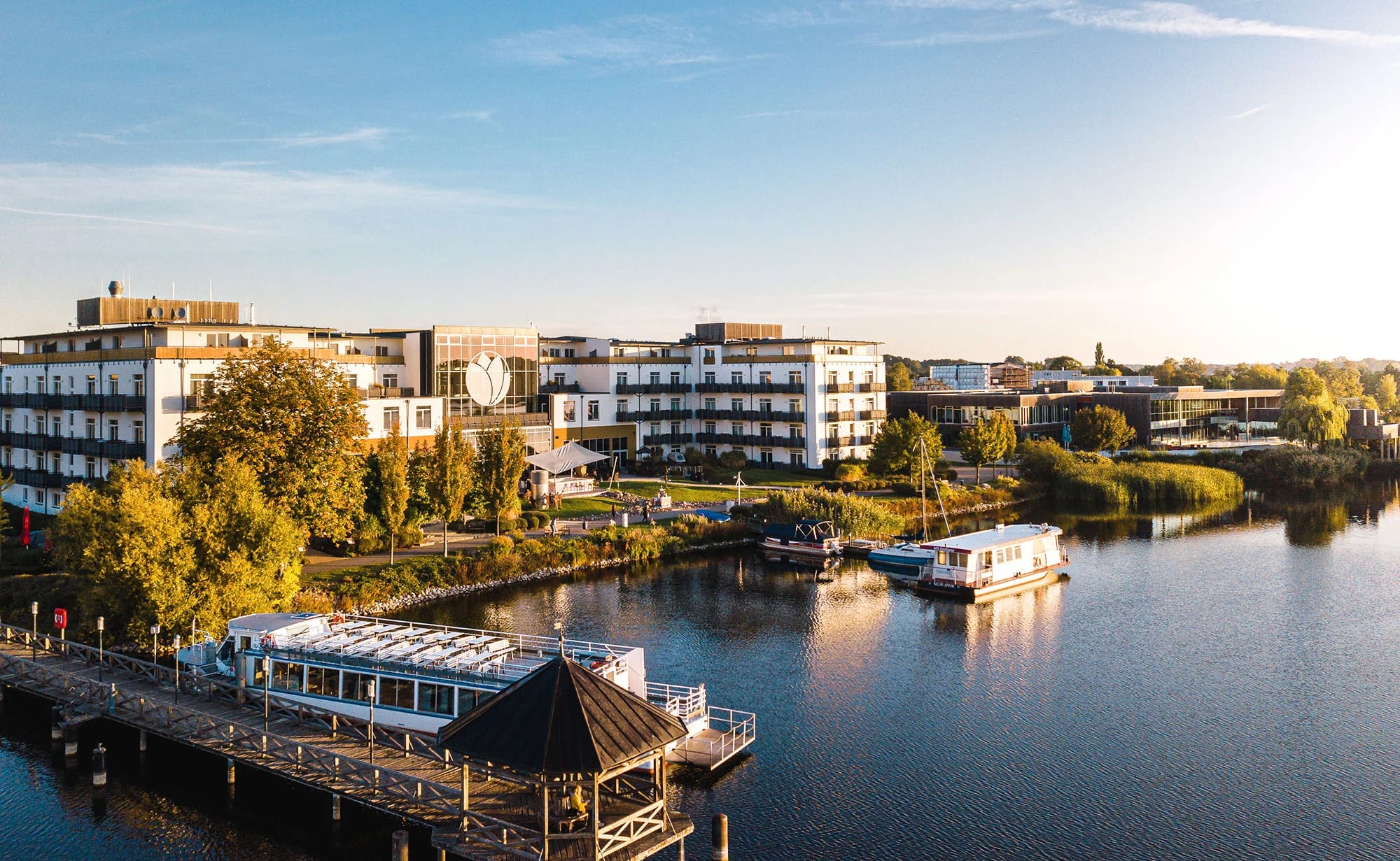 Blick über den Ruppiner See auf die langgestreckten Gebäude des Resorts im Abendlicht, davor ein Steg und ein Ausflugsschiff