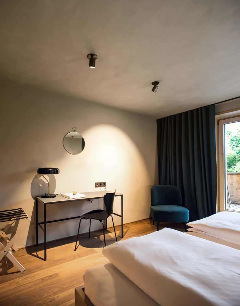 Zimmer mit Schreibtisch, rundem Spiegel, blauem Sessel und Bett vor der Terrassentür